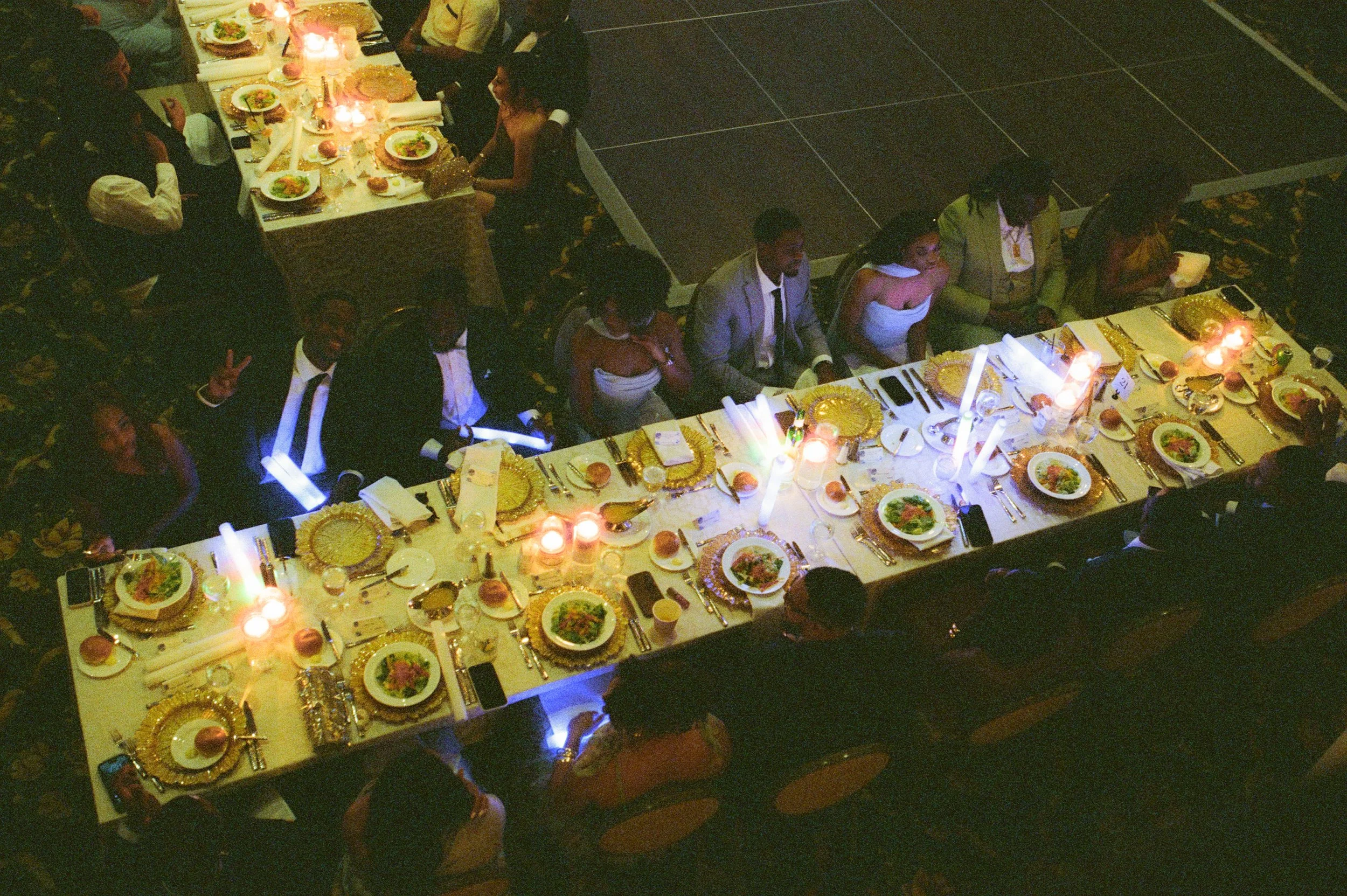 Overhead view of guests seated at long reception tables with glowing candles and elegant gold place settings captured in cinematic film photography style