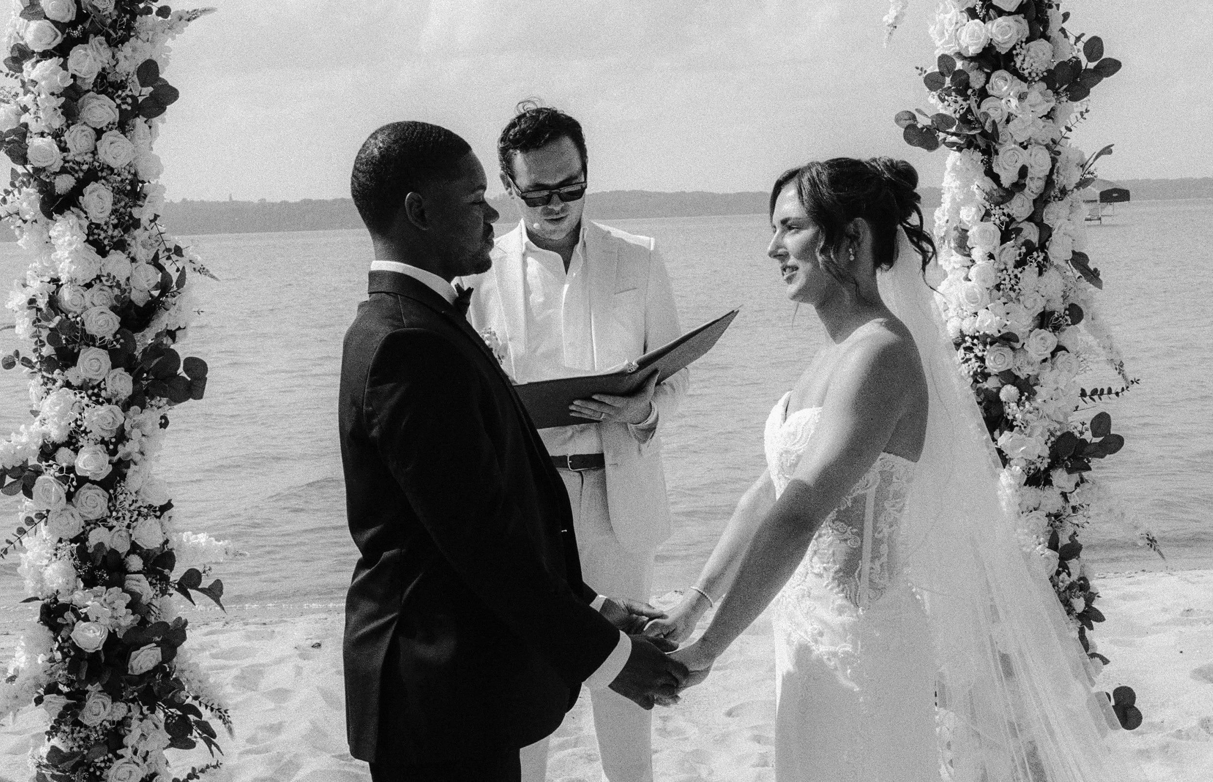 Black and white photo of the bride and groom holding hands and exchanging vows beside the lake.