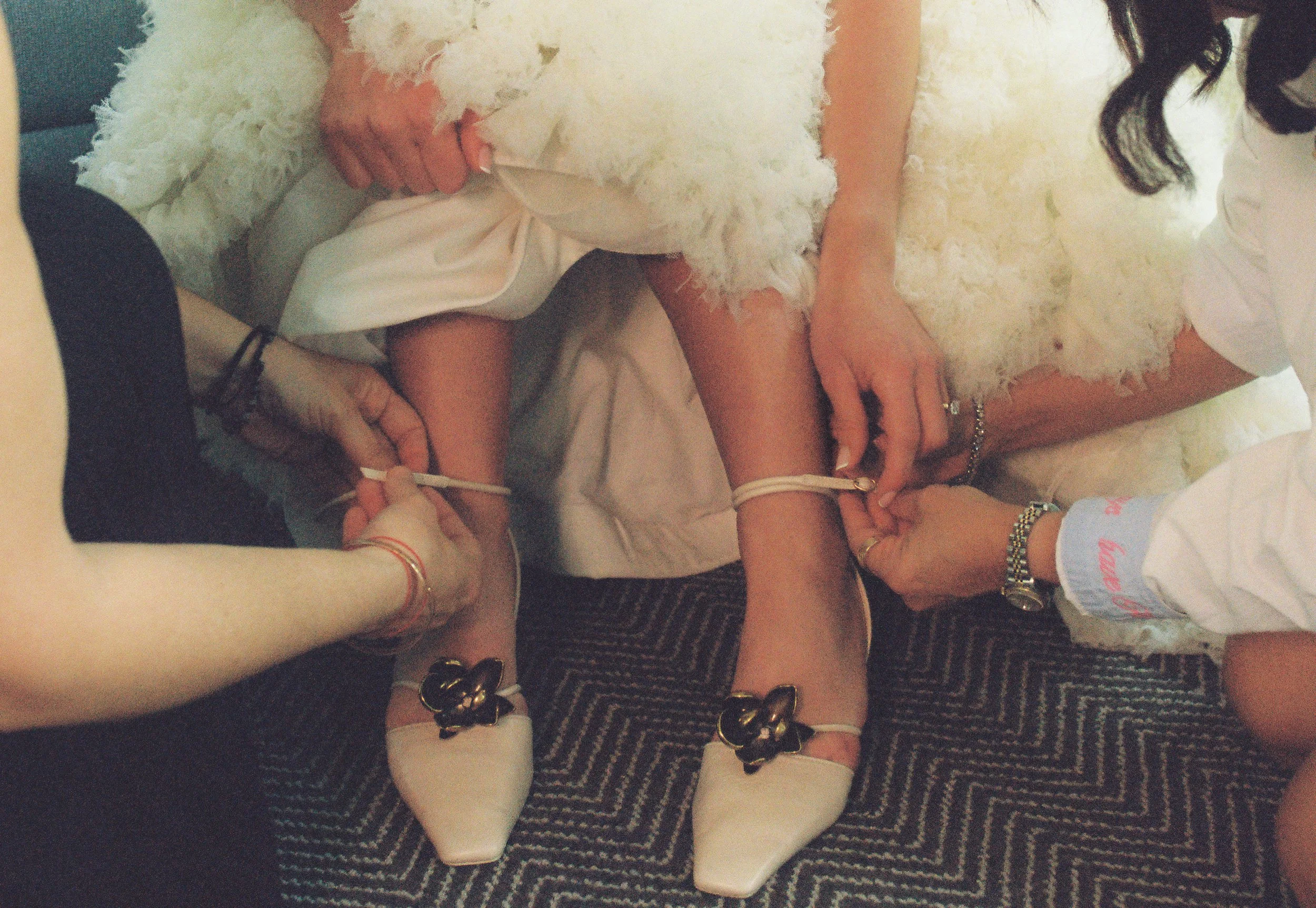 Bridesmaids helping fasten the bride’s ankle-strap wedding shoes during getting ready moments.