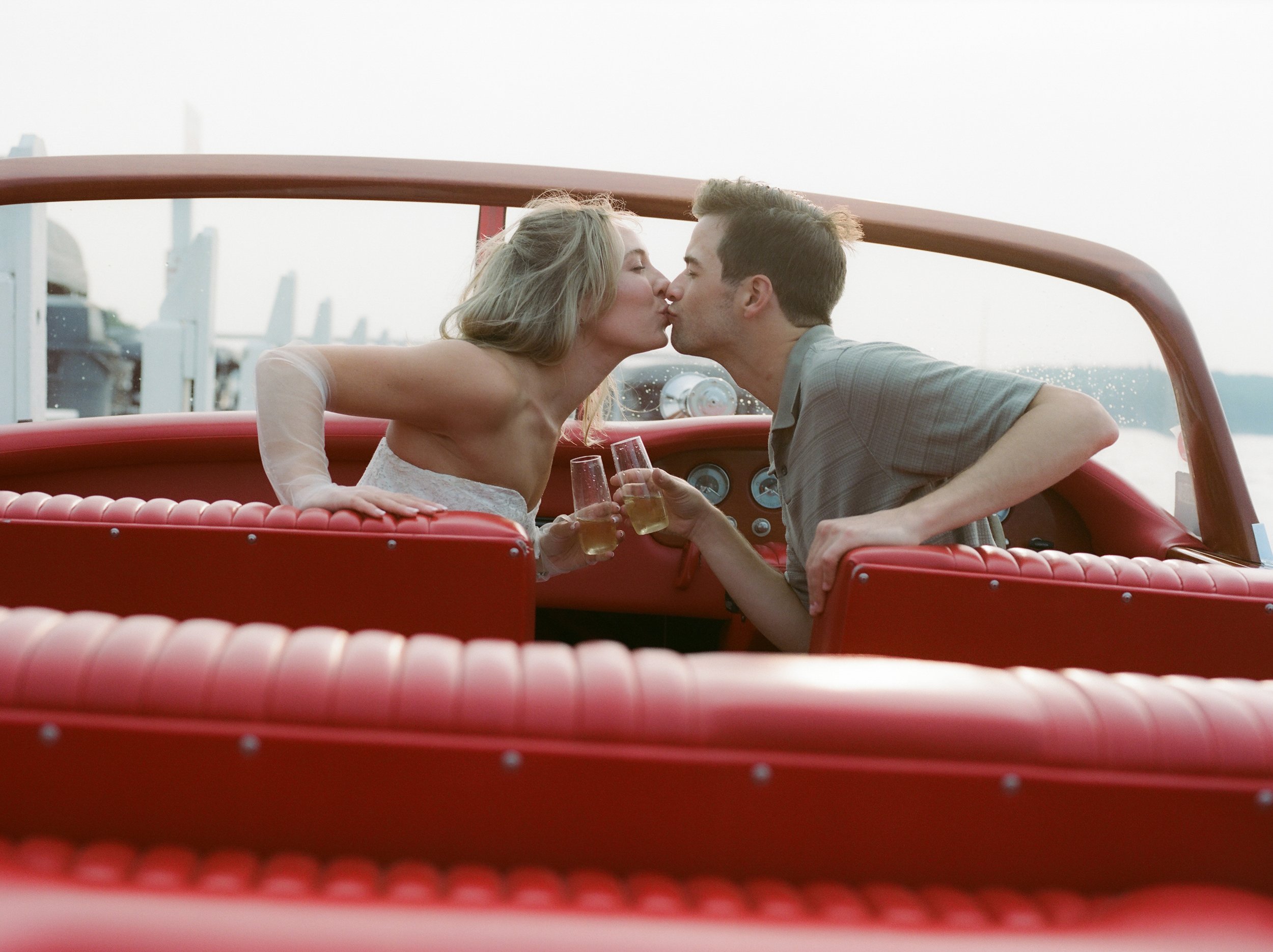 Couple sharing a kiss while toasting with champagne aboard a vintage wooden boat during a Lake Geneva Engagement session at sunset.