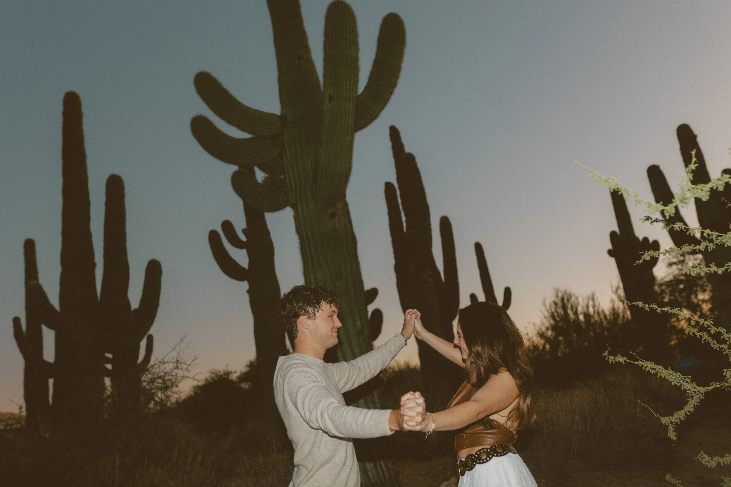 Couple holding hands and spinning together among tall cacti at dusk, captured with flash for a fun and energetic couple's photoshoot.