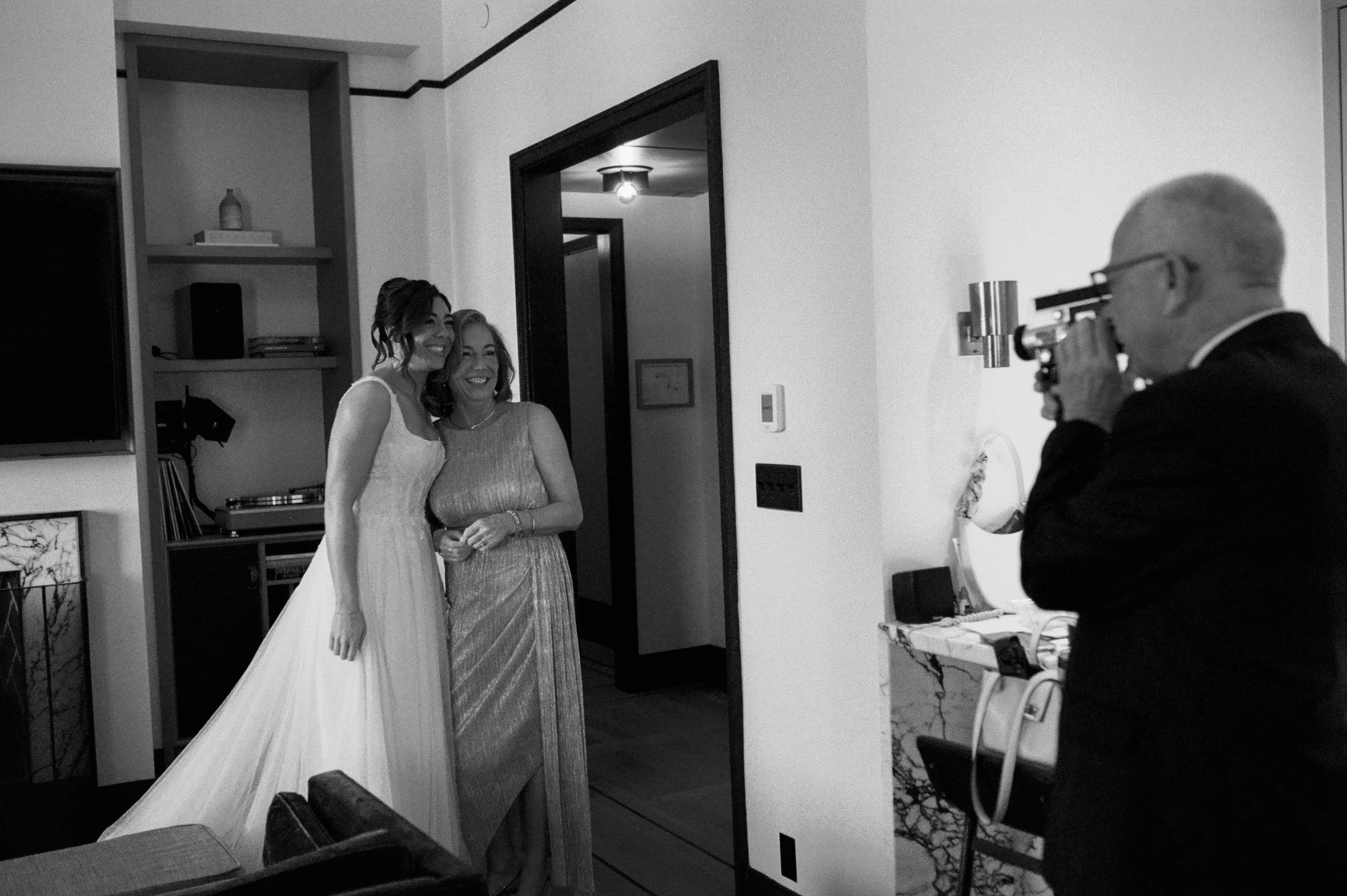 Black and white photo of the bride posing with her mother while a family member takes a photo inside the hotel suite