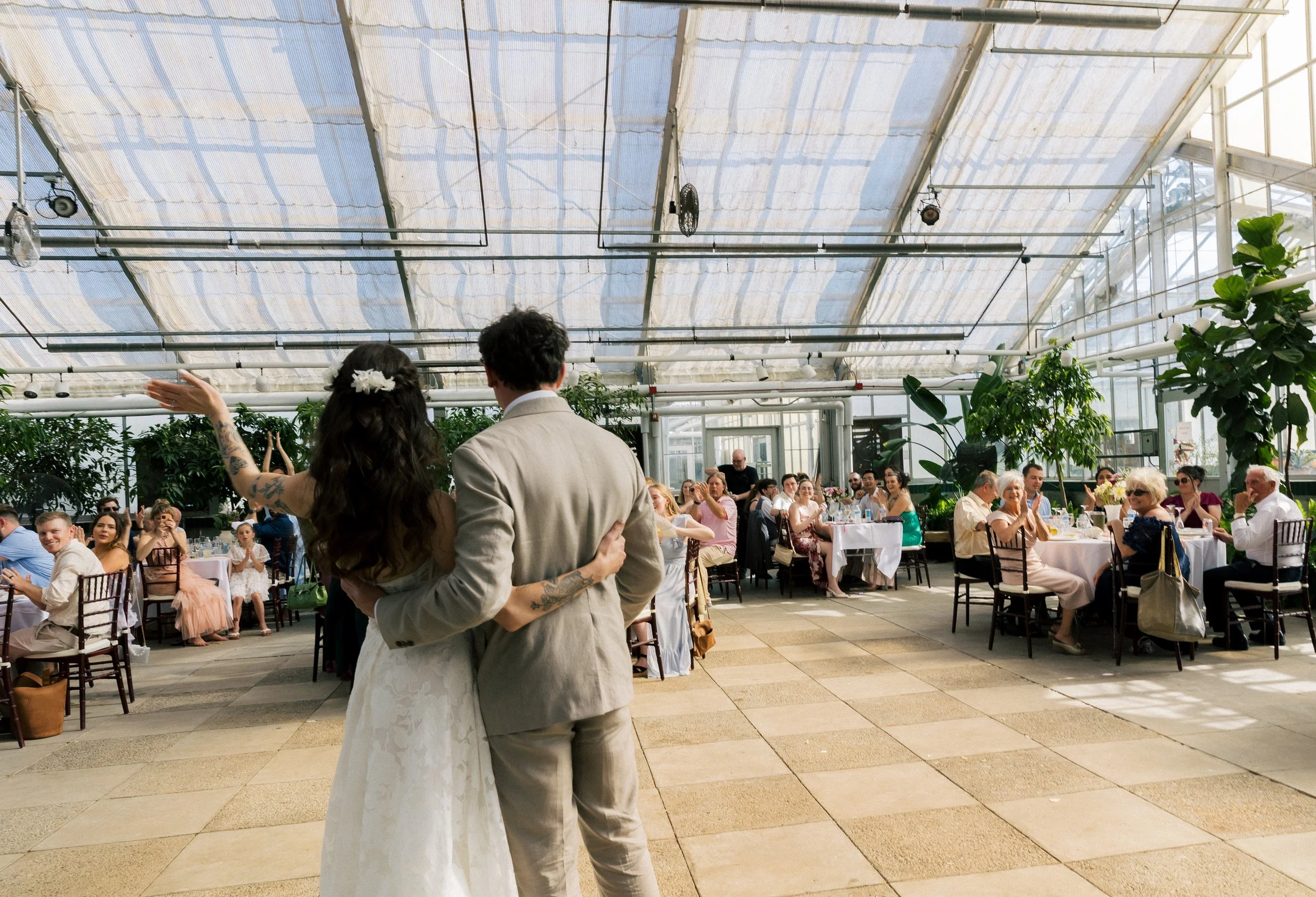 Bride and groom standing arm in arm facing their seated guests during the reception inside a greenhouse