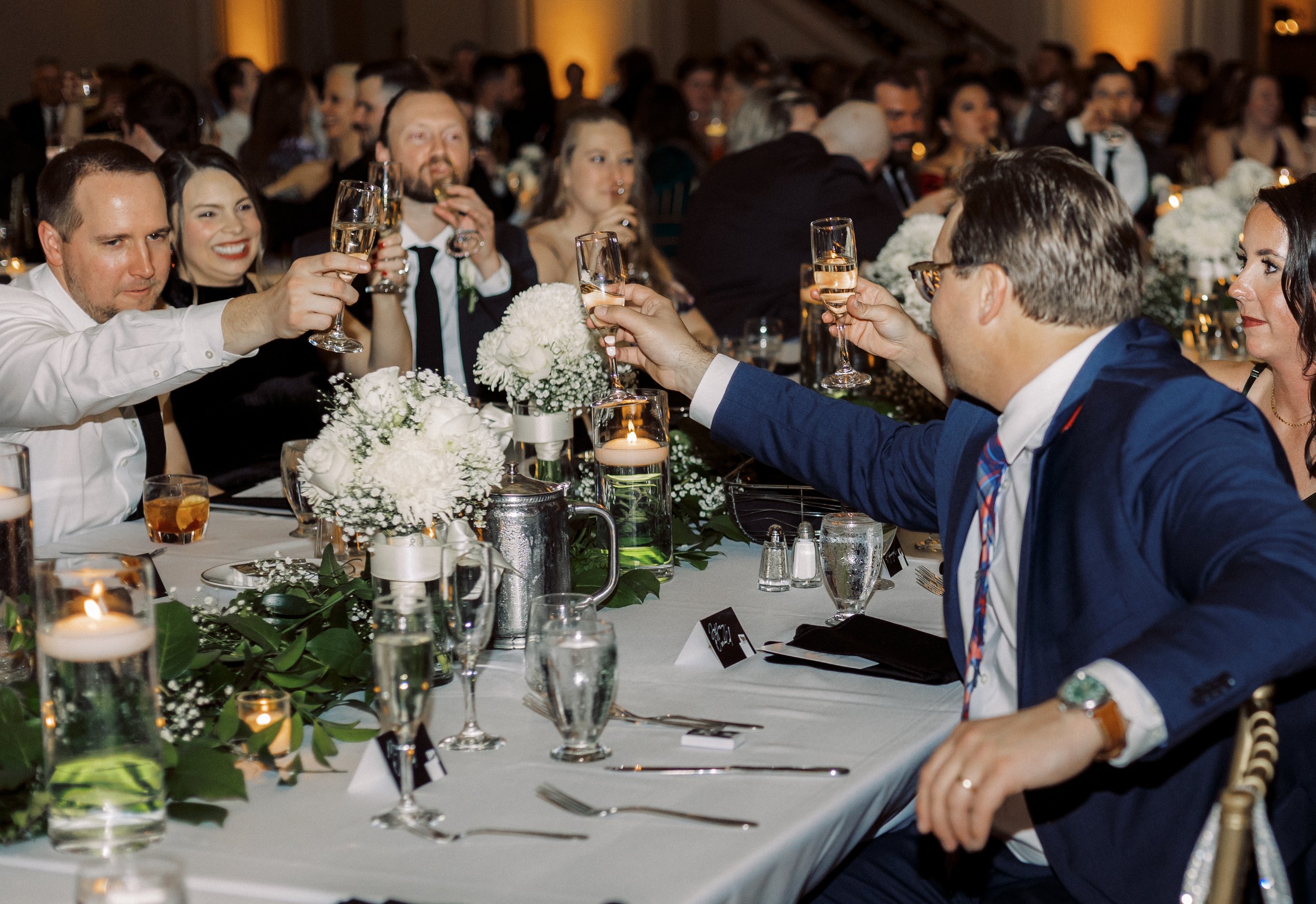 Wedding guests raising champagne glasses for a toast around a reception table decorated with white flowers and greenery.