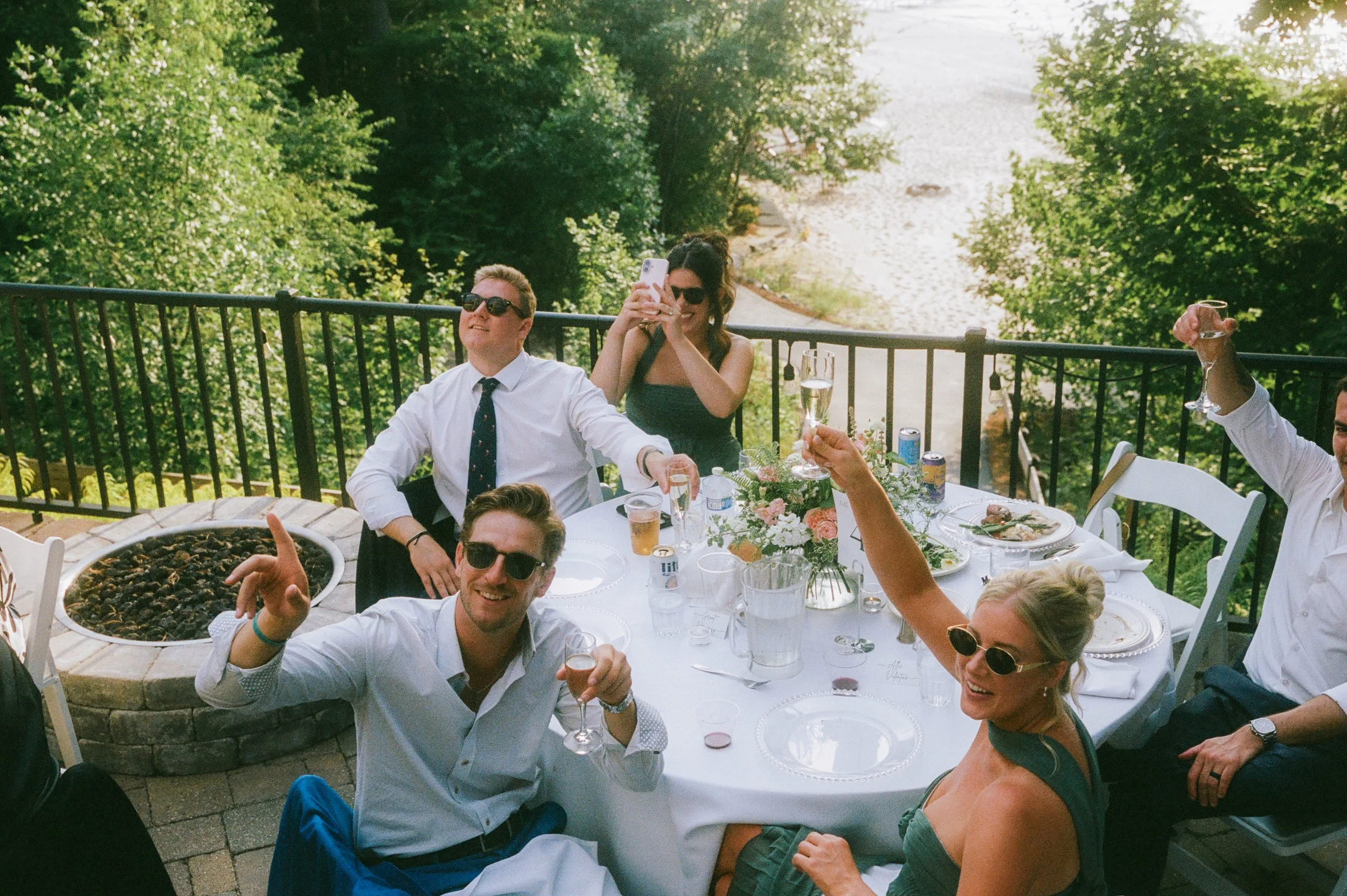 Wedding guests raising champagne glasses around an outdoor reception table overlooking the water.