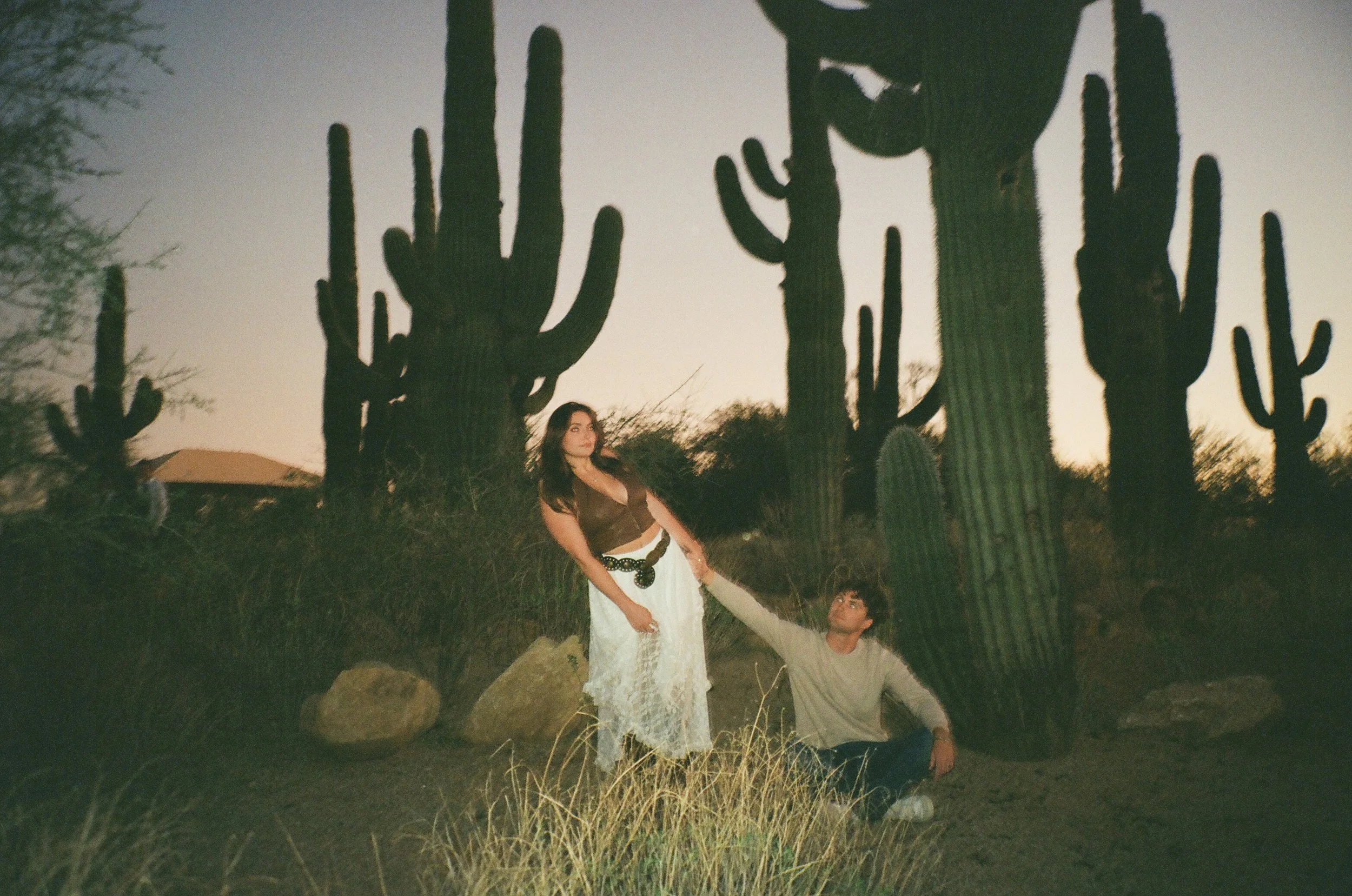 Playful scene of a couple interacting among tall cacti at dusk with flash lighting, adding a nostalgic and artistic feel to the couple's photoshoot.
