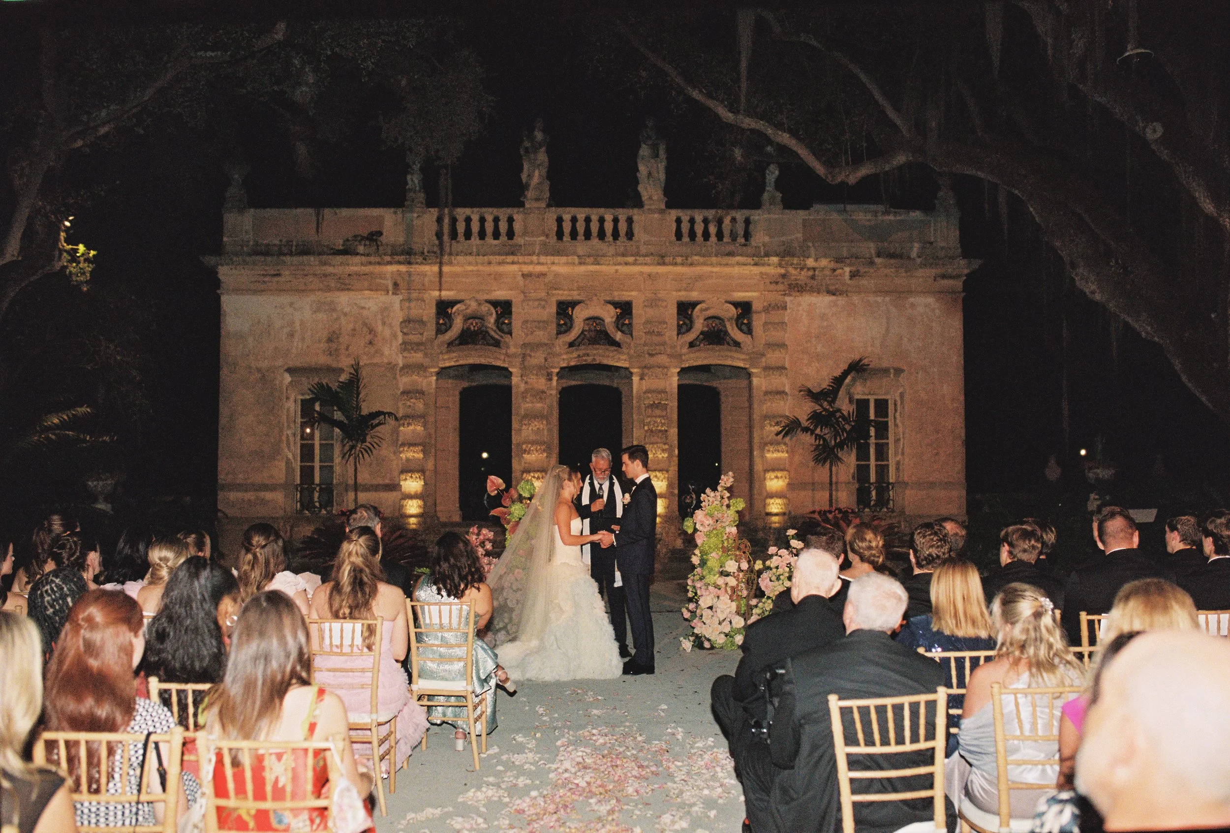 Outdoor wedding ceremony at night with guests seated and floral arrangements framing the altar at Vizcaya Museum and Gardens.