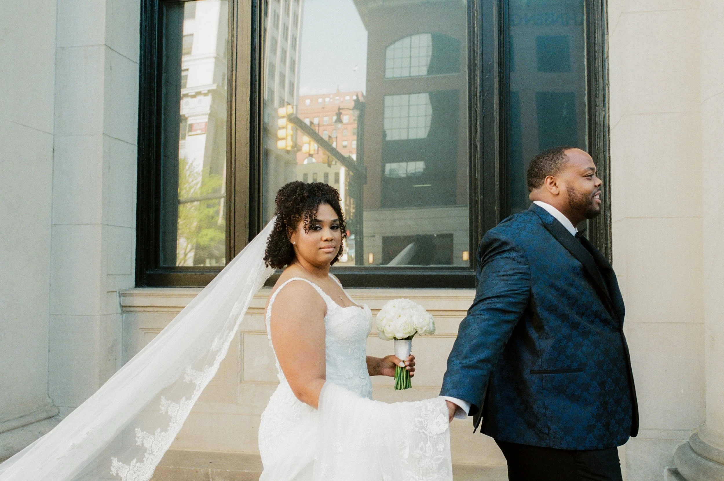 Bride holding a bouquet and lace veil while walking hand in hand with the groom outside their wedding venue