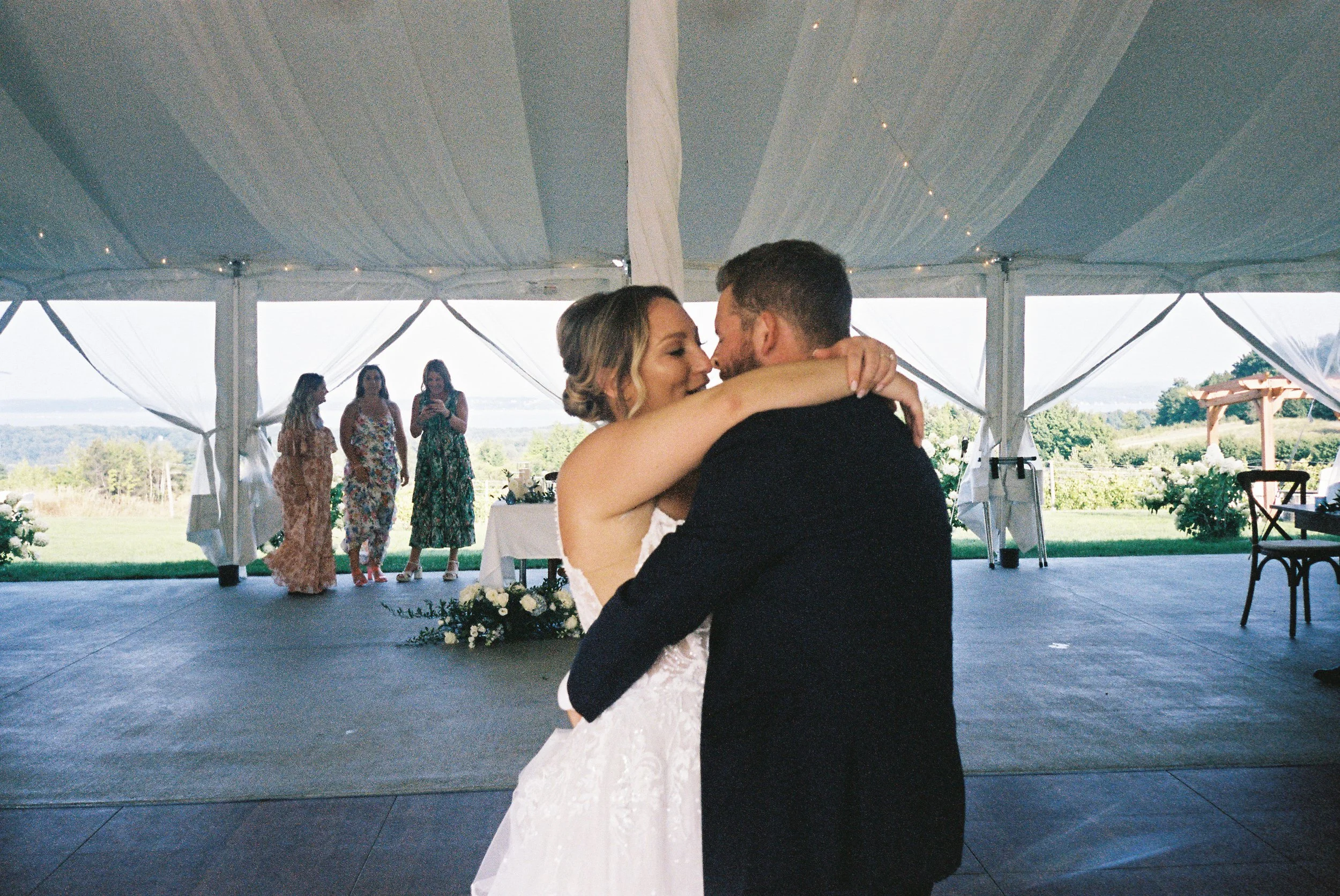 Newlywed couple sharing their first dance beneath a white reception tent surrounded by family and friends.