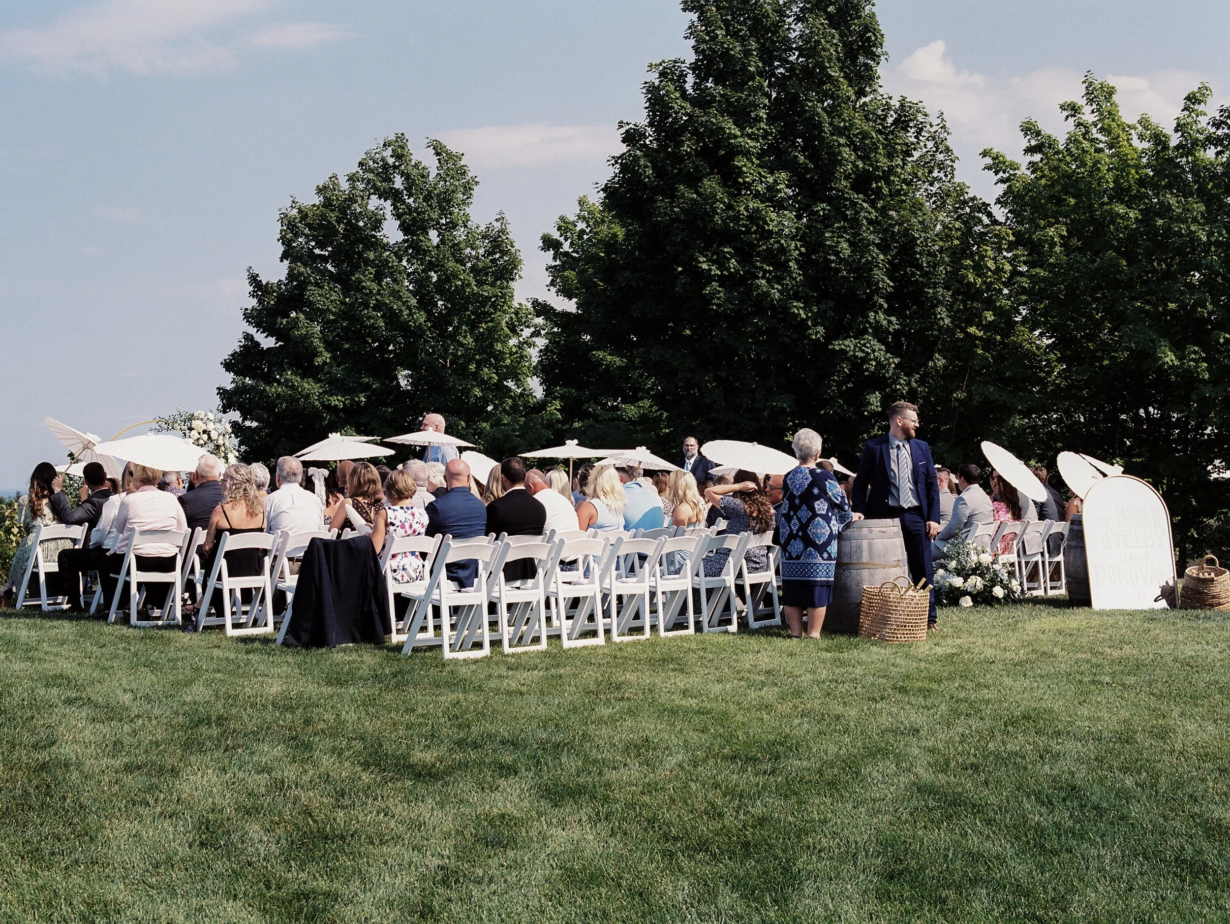 Outdoor wedding ceremony with guests seated on white chairs beneath parasols overlooking a vineyard landscape.