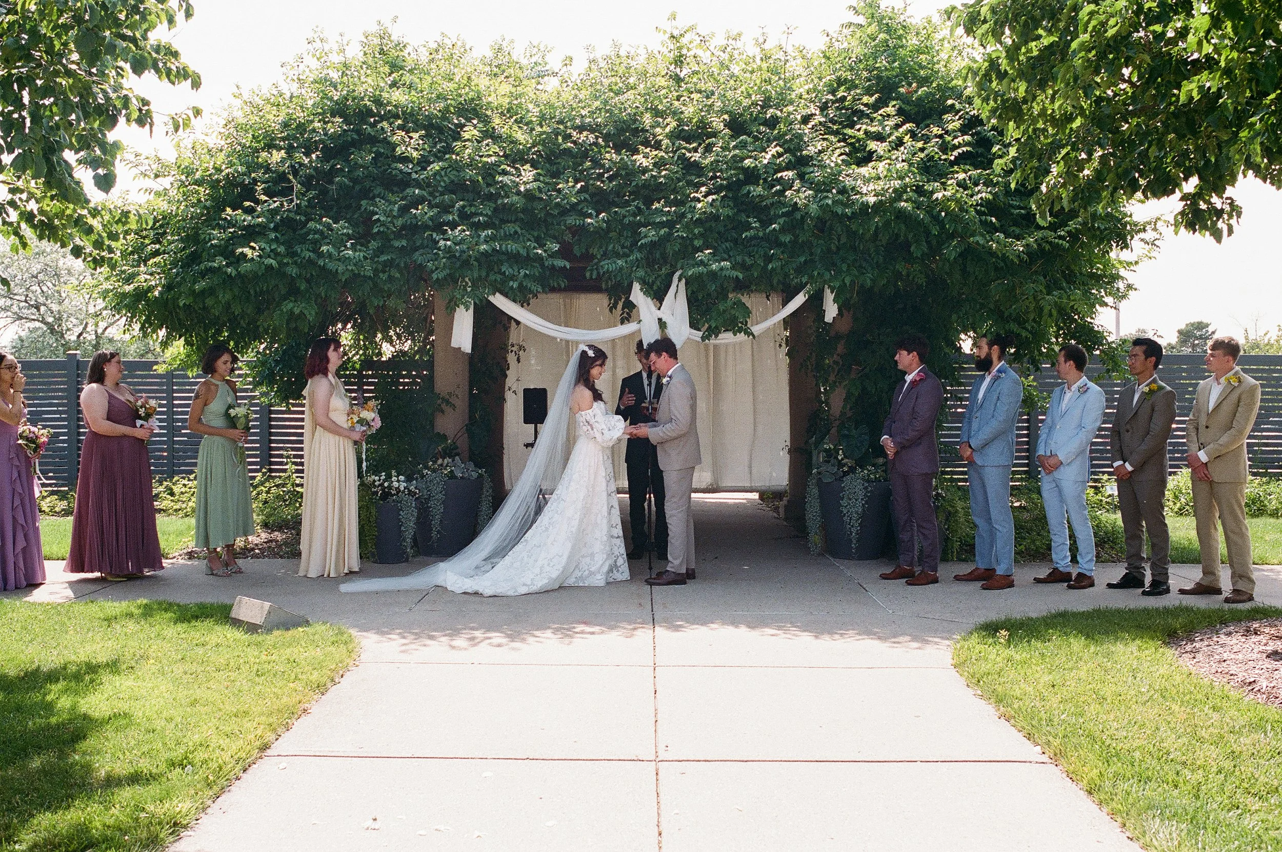 Bride and groom exchanging vows during an outdoor ceremony beneath a leafy arbor with wedding party standing nearby