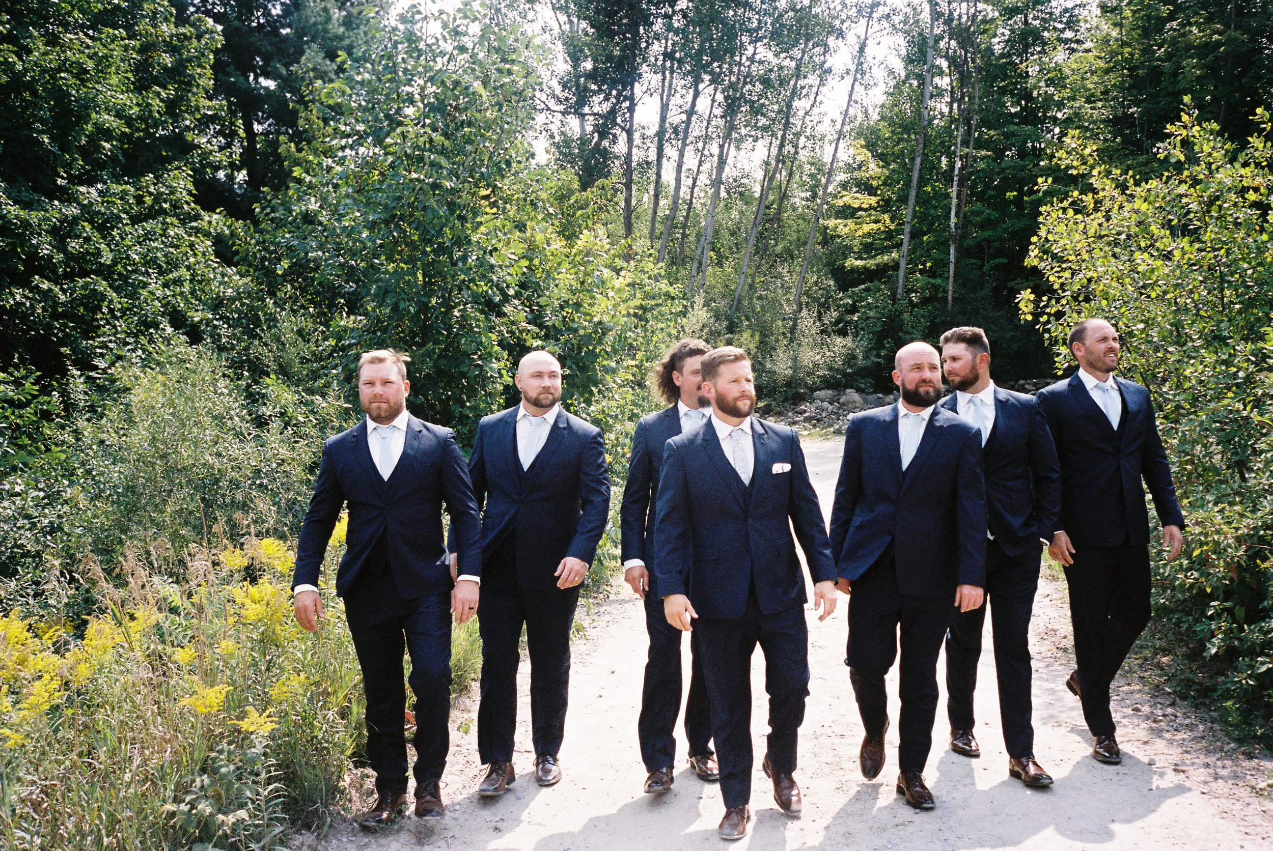 Groom and groomsmen walking together down a tree-lined path before a Northern Michigan vineyard wedding celebration.