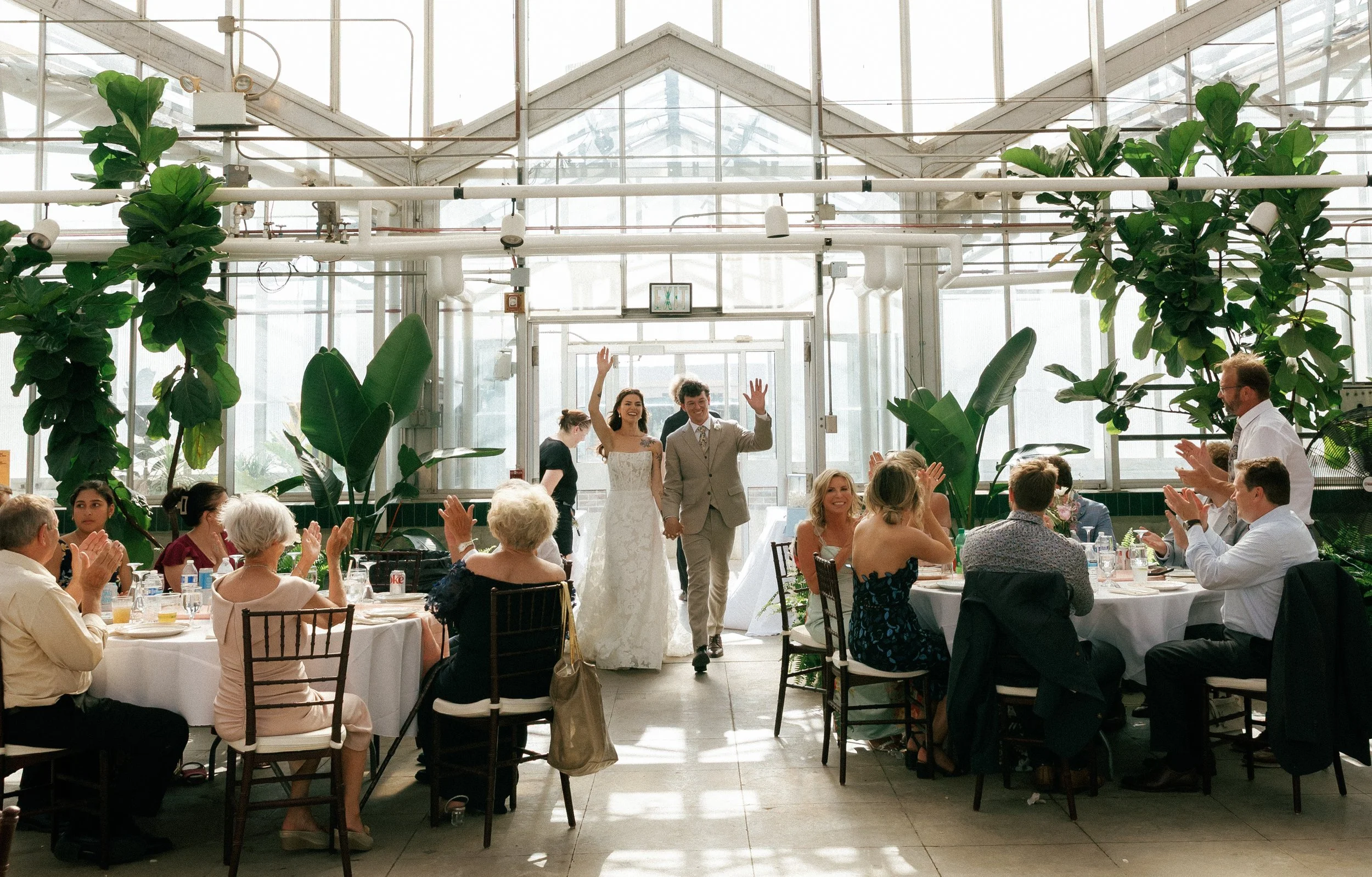 Newlyweds walking into their reception while guests clap and celebrate, captured in a bright film wedding photography style
