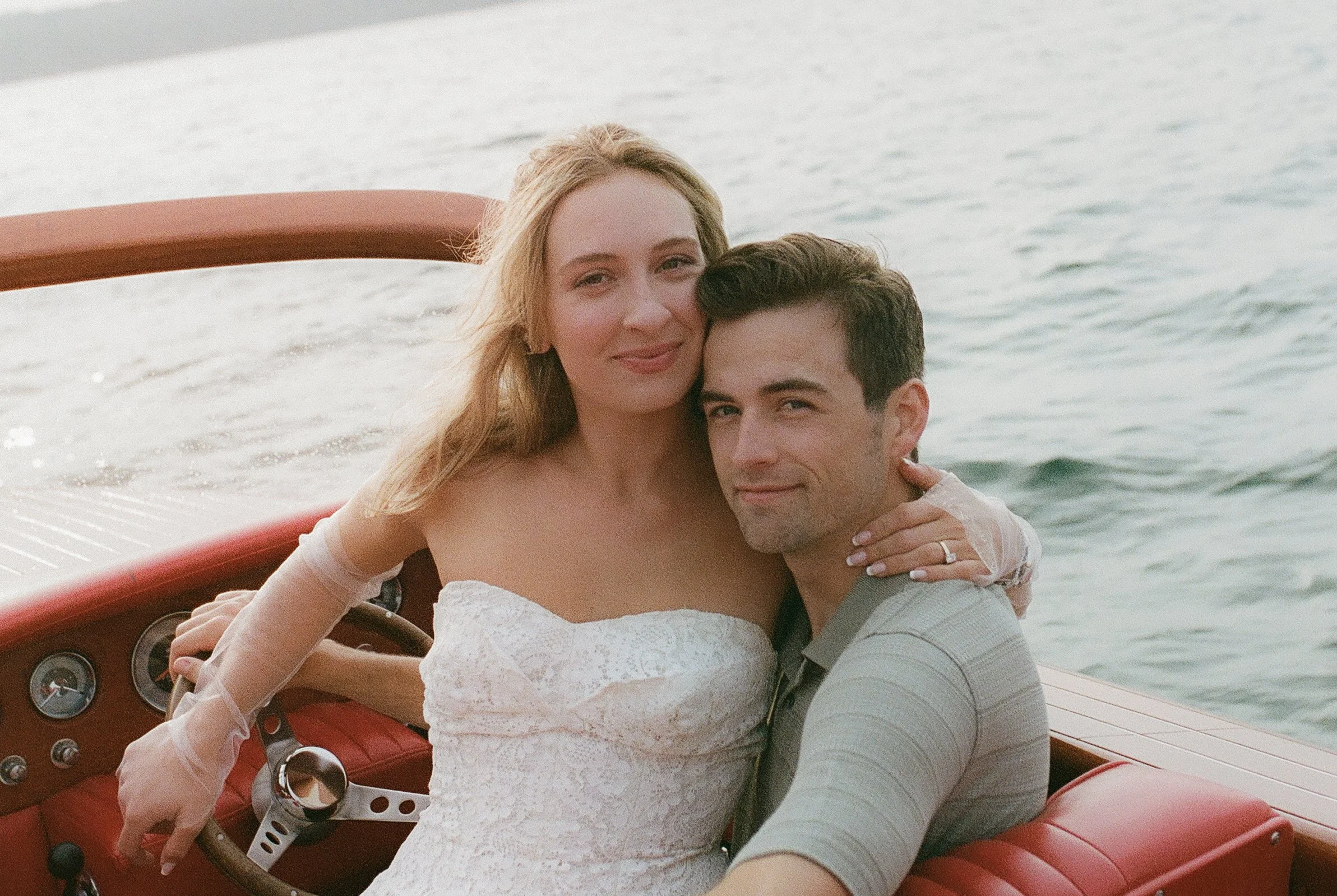 Close-up portrait of an engaged couple embracing aboard a wooden boat with soft golden evening light reflecting off the water.