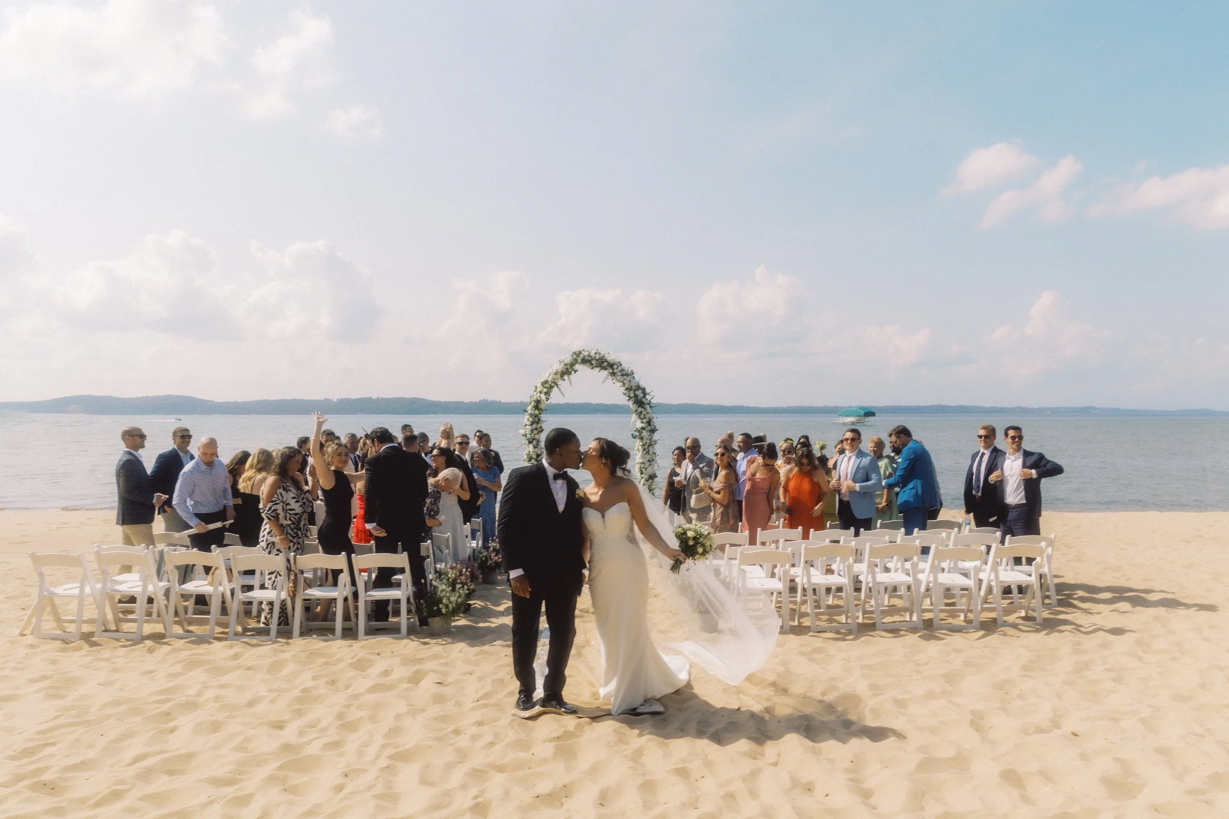 Bride and groom kissing on the beach as guests celebrate behind them after their lakeside ceremony.