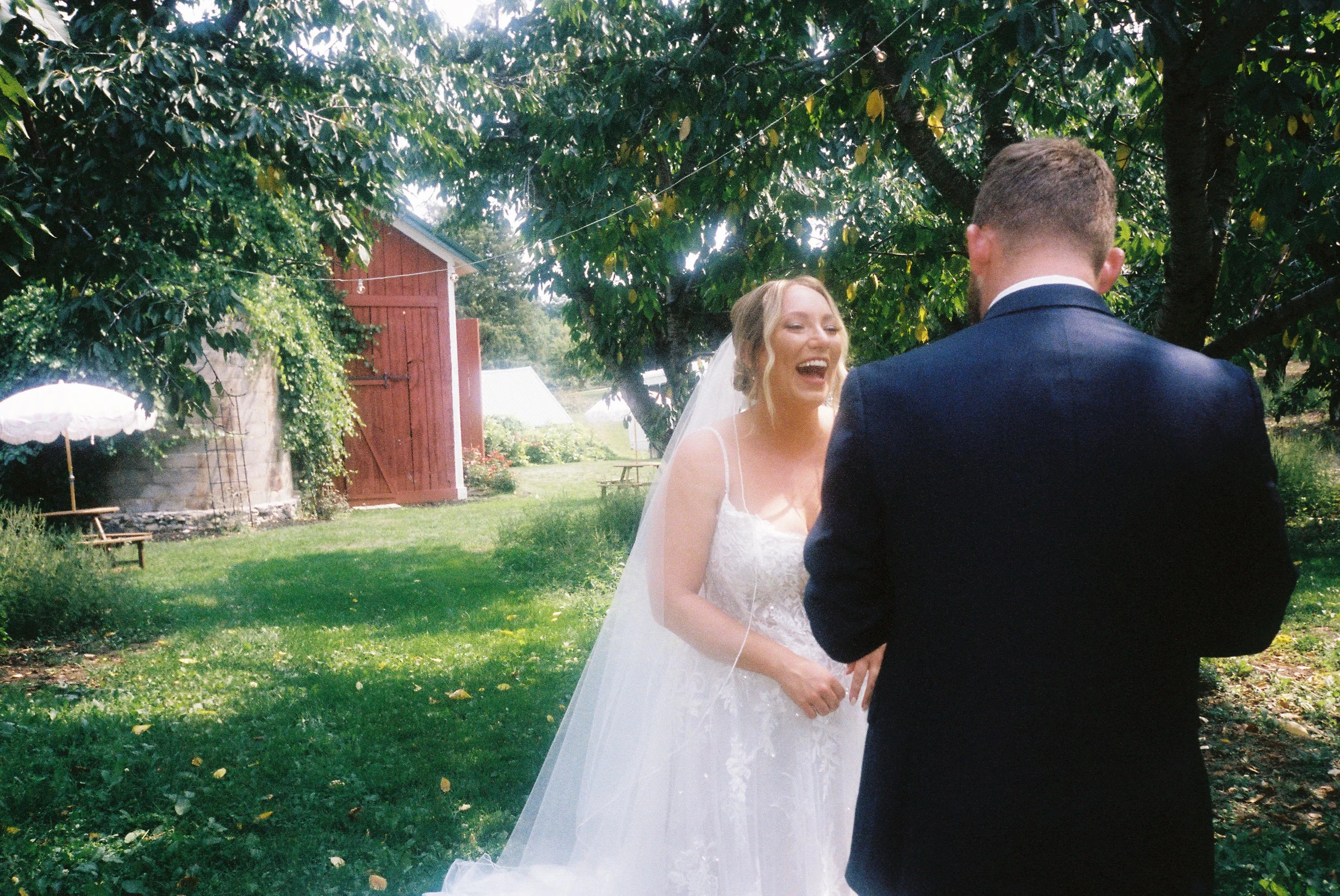 Bride laughing during a first look with her groom beneath trees near a rustic red barn wedding venue.