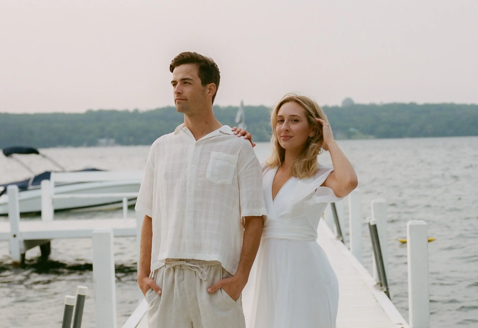 Bride resting her hand on her fiancé's shoulder while posing together on a dock overlooking the lake.
