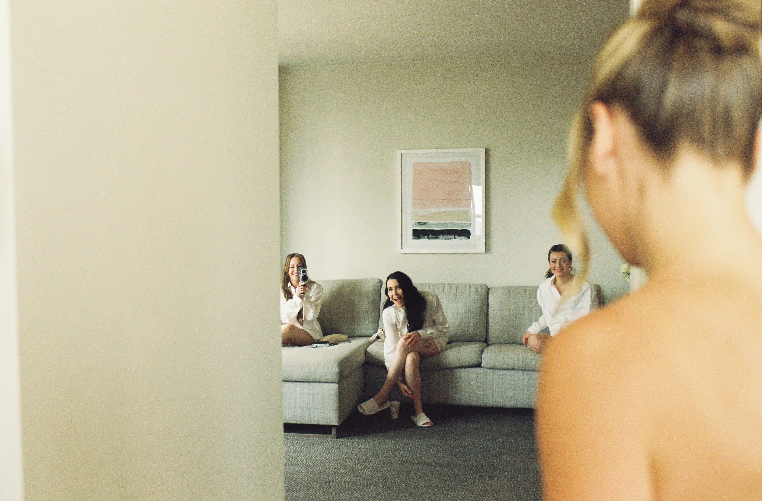 Bridesmaids sitting on a couch laughing while watching the bride get ready for her wedding day.