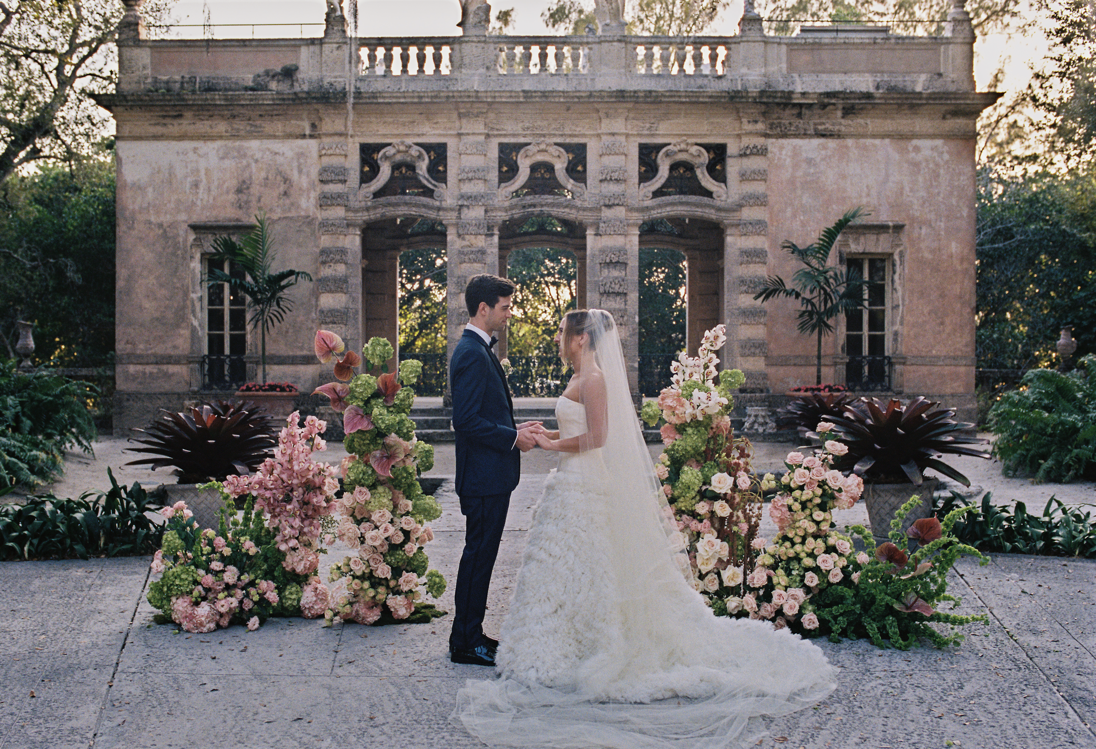 Bride and groom standing together during their ceremony surrounded by lush floral arrangements in the courtyard.