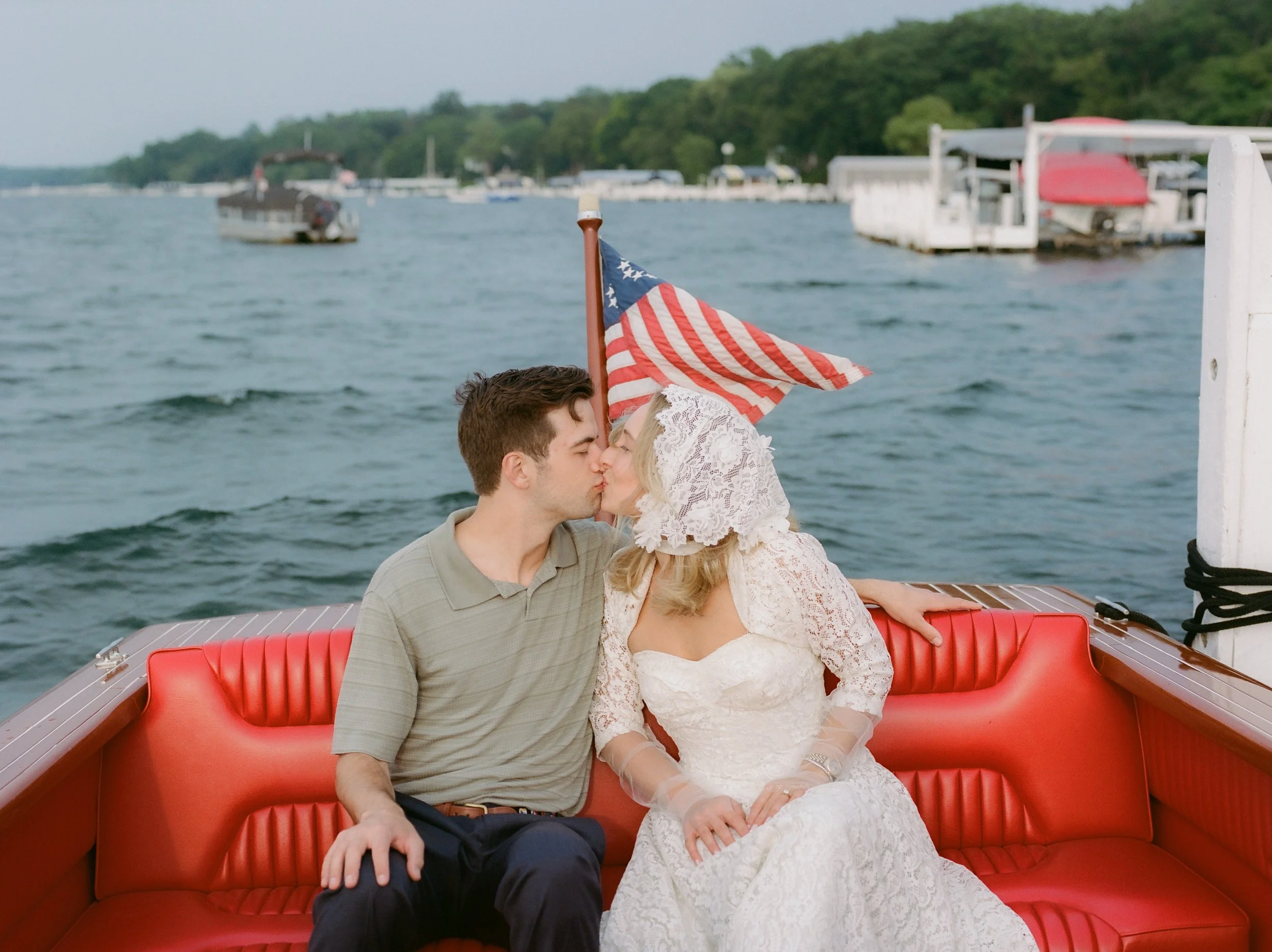 Bride and groom sharing a kiss aboard a classic wooden boat with an American flag waving in the background during their Lake Geneva Engagement session.