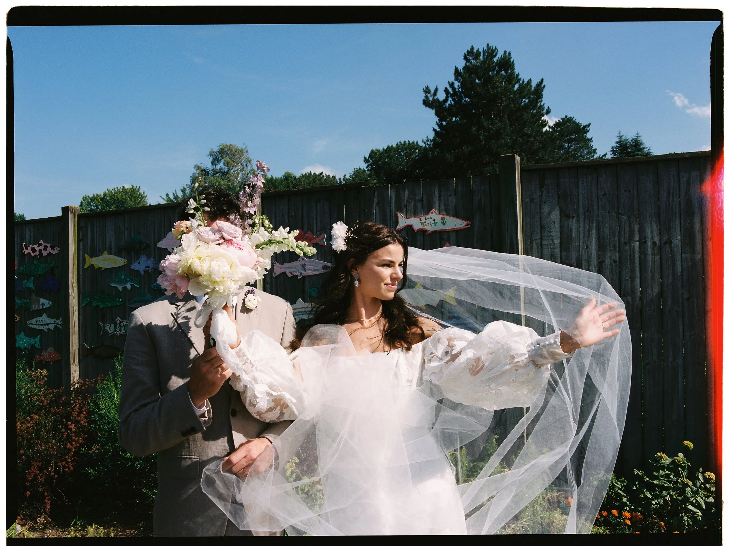 Bride holding out her veil in the sunlight while groom stands beside her holding the bouquet, captured in a playful film wedding photography style