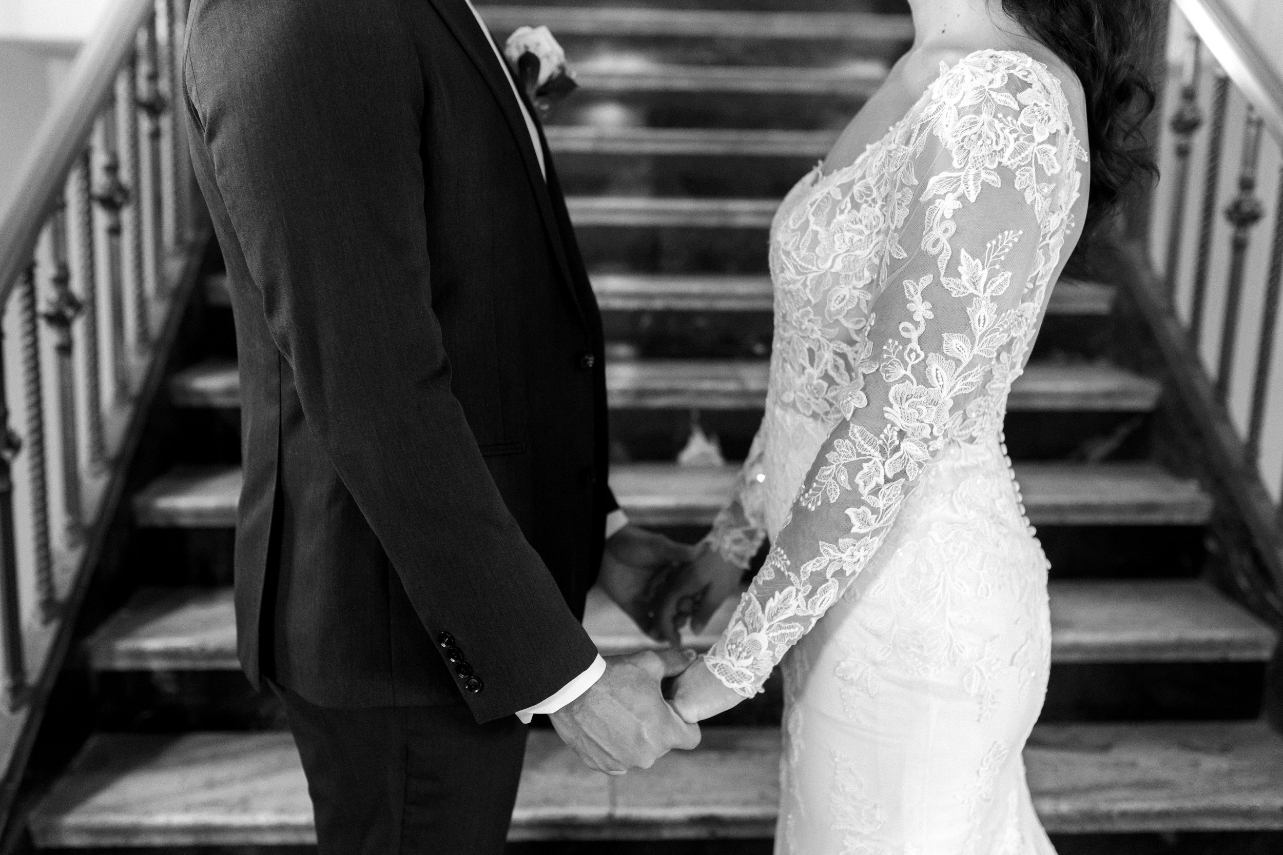 A black and white image of a close up of a bride and groom holding hands sharing the details of the brides lace wedding dress