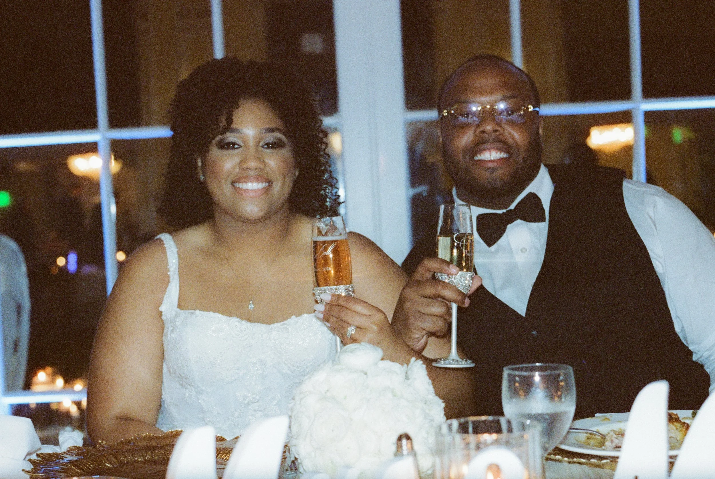 Bride and groom smiling during a champagne toast at their wedding reception with glowing candlelight and cinematic film photography tones