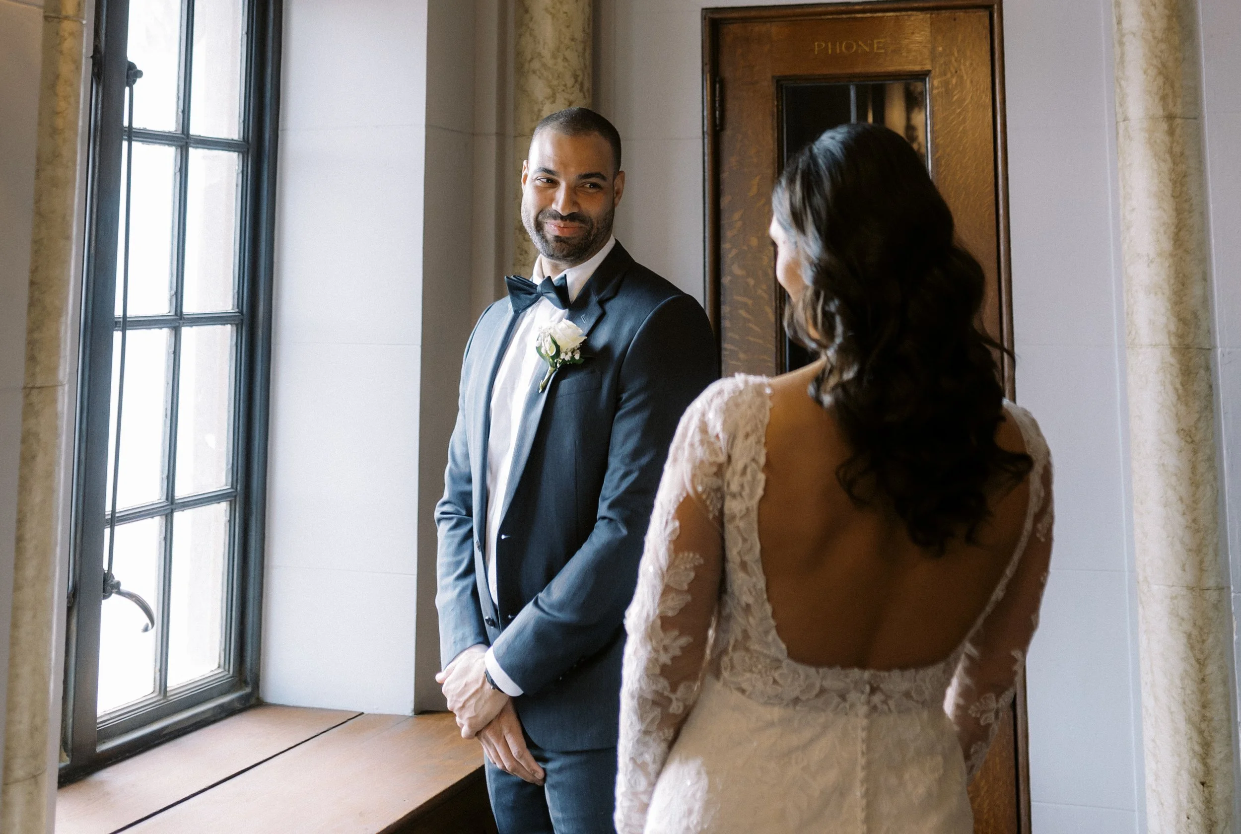Groom smiling at the bride during an intimate first look beside a large window.