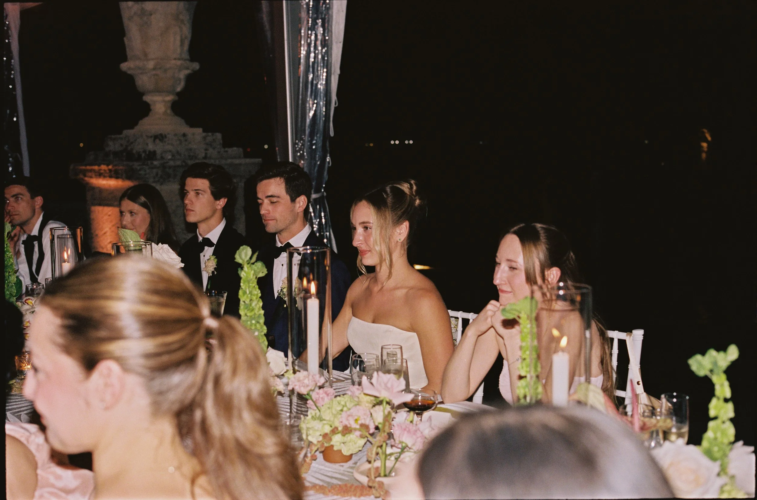 Bride and groom seated at a candlelit reception table surrounded by guests and soft floral arrangements during their Vizcaya Museum and Gardens wedding reception.