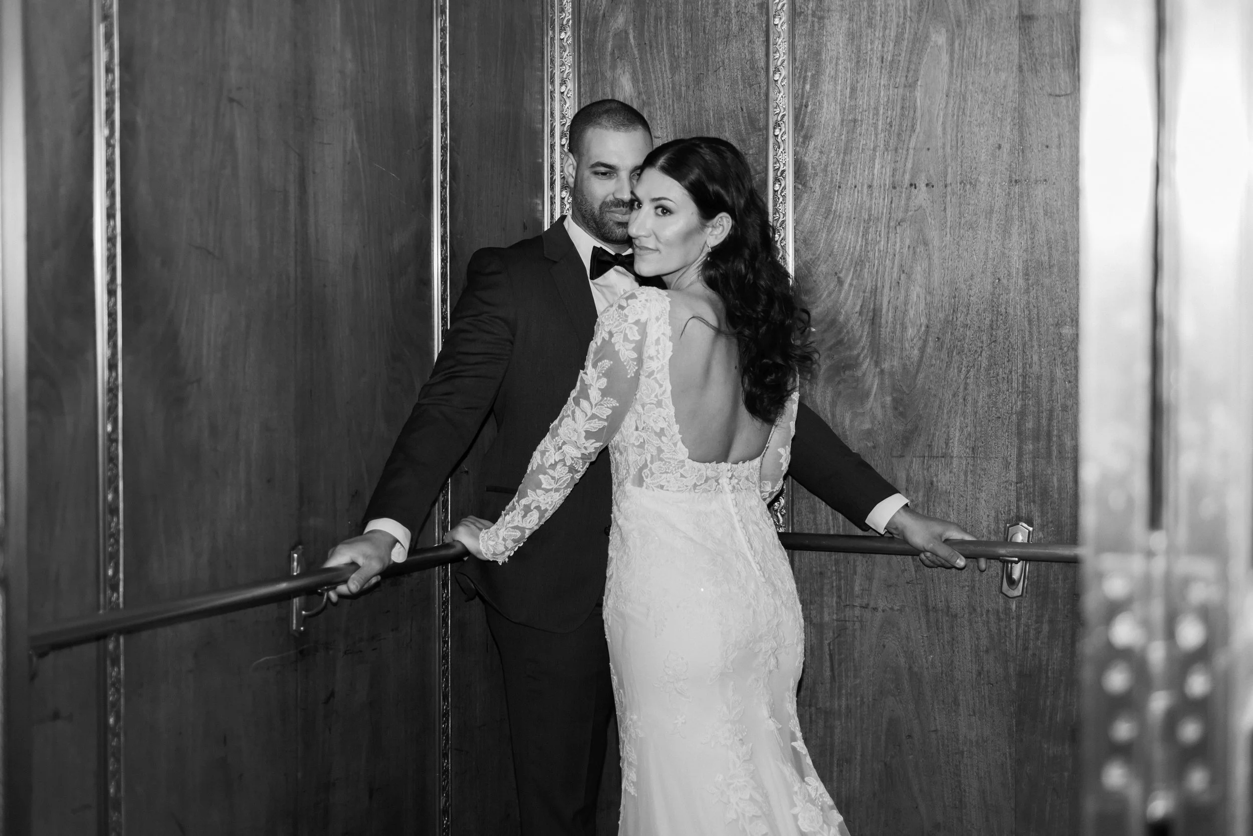 A black and white image of a bride and groom in an elevator, the bride looking back a the camera