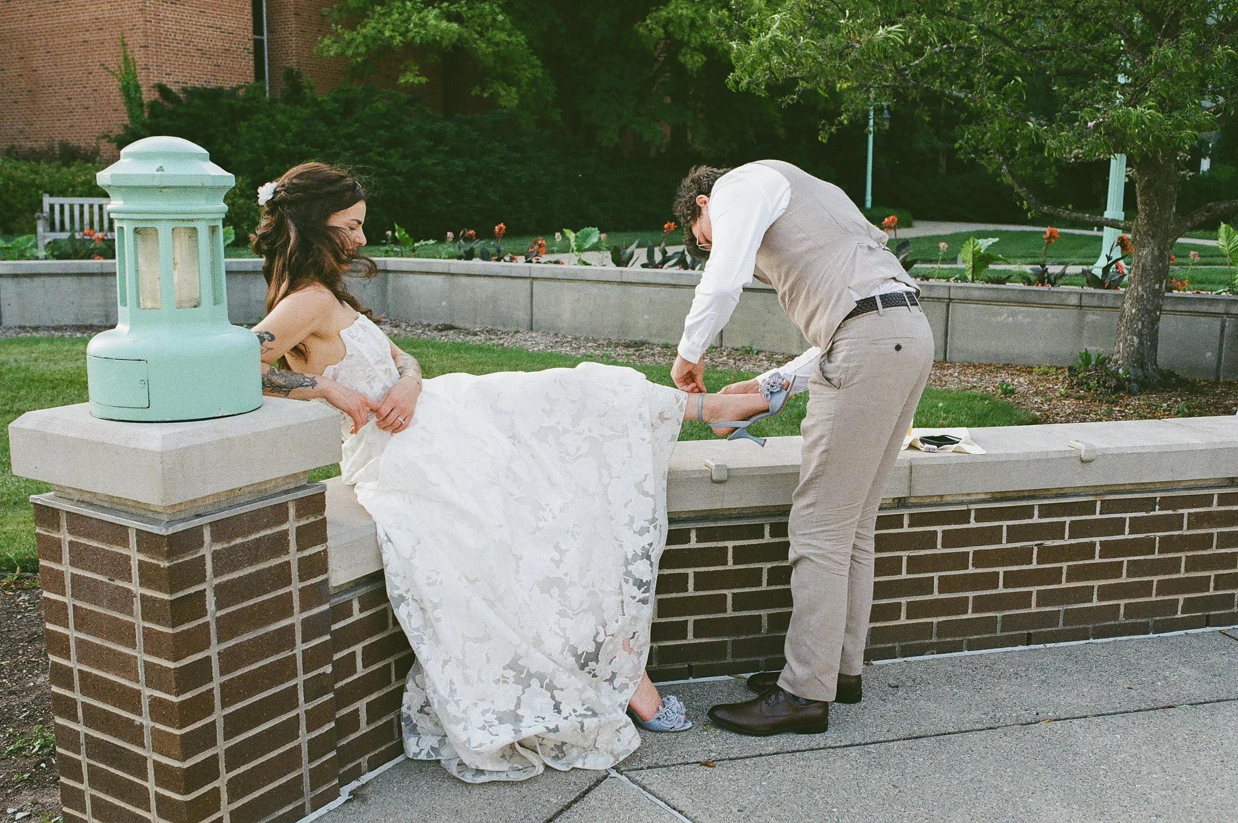 A bride sitting on a brick ledge while her groom is adjusting her blue wedding shoes.