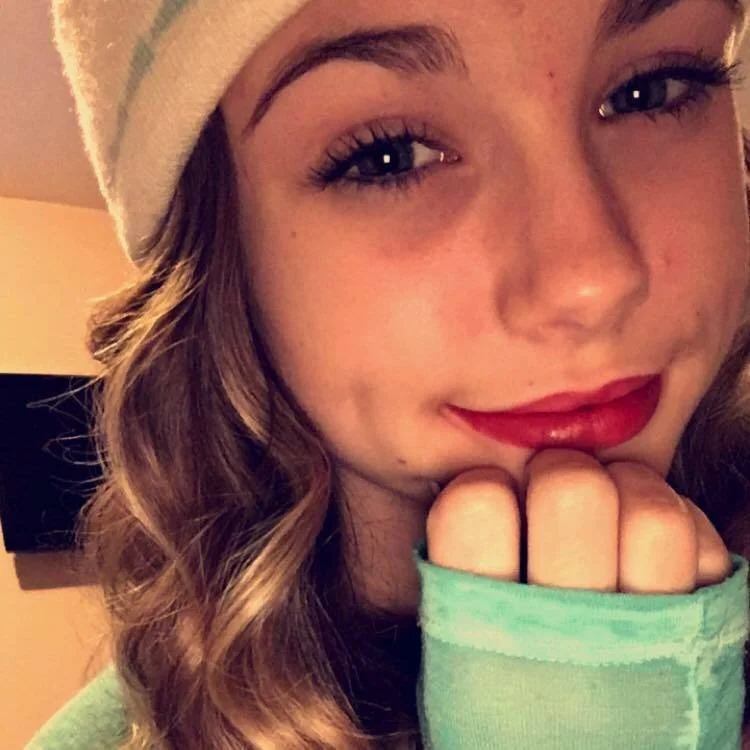 Close-up of a young woman with wavy hair wearing a beanie, smiling slightly with her chin resting on her hand, in a cozy indoor setting.