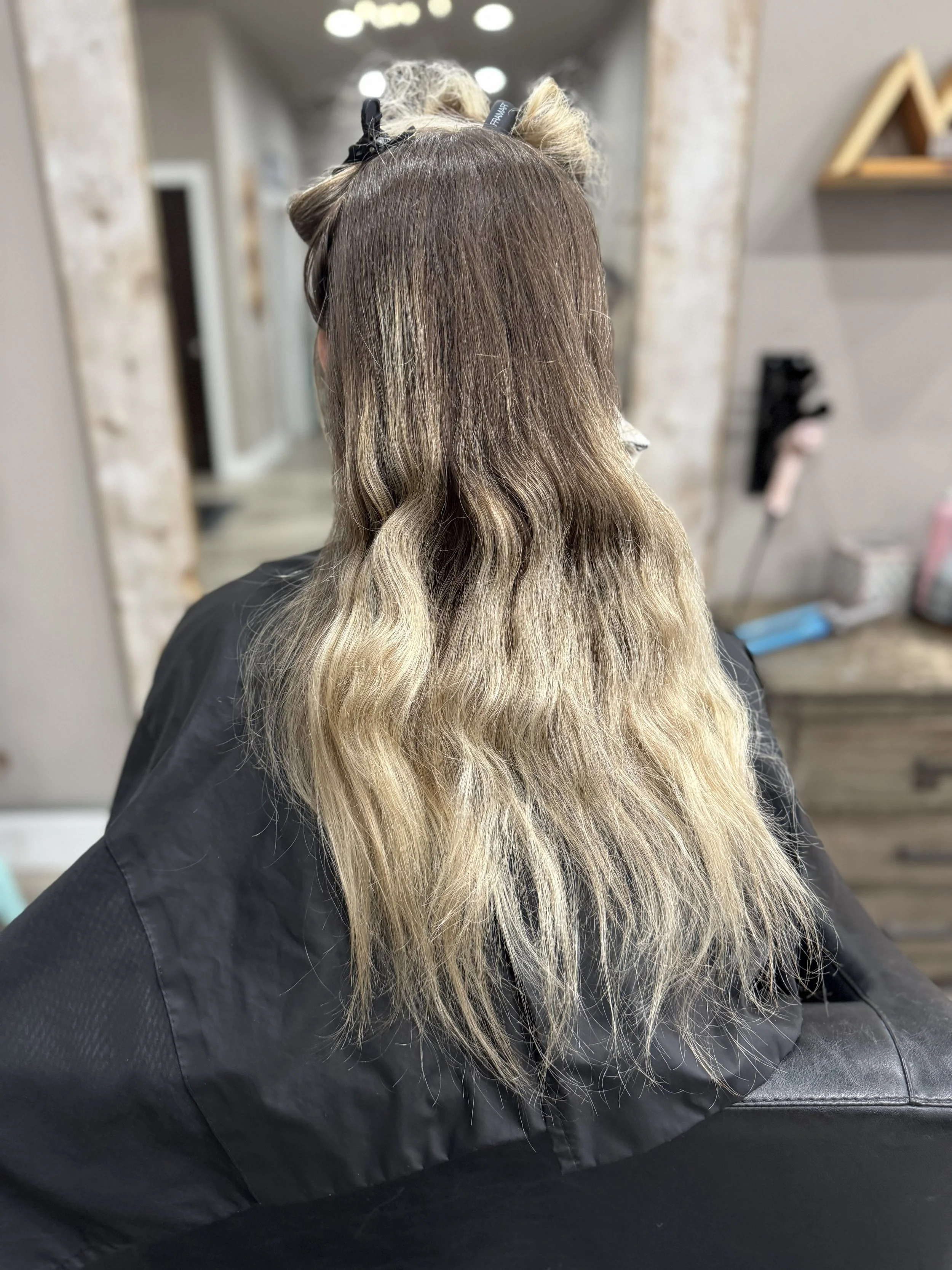 Before the transformation, back view of a woman with shoulder-length wavy hair, cut in a style with darker roots and light blonde ends, sitting in a hair salon.