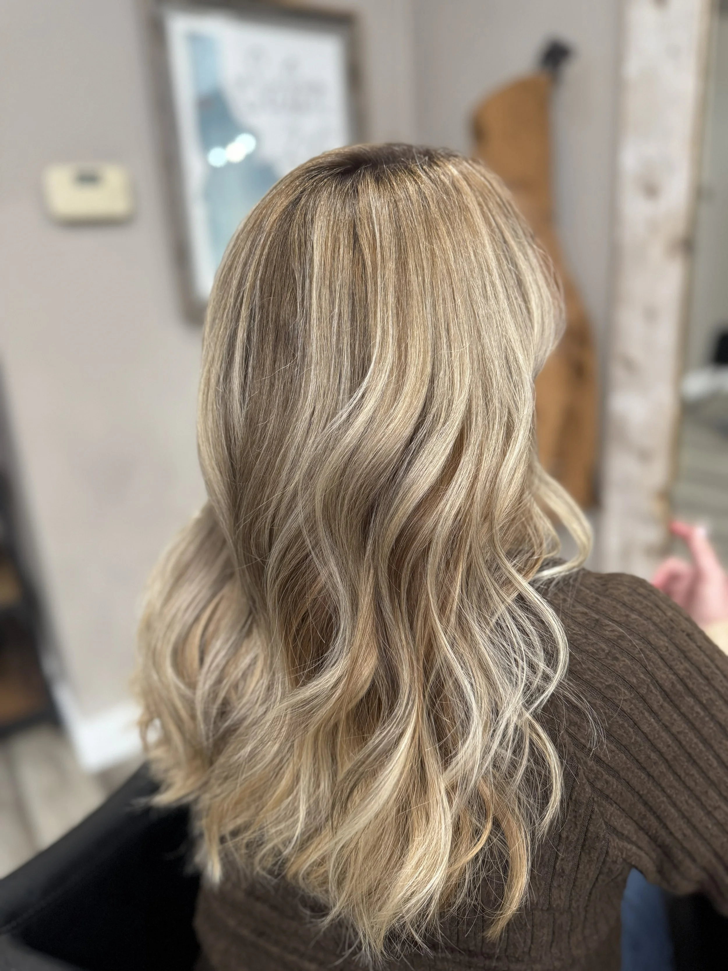 After the transformation, a woman's blonde, wavy hair is viewed from the back in a salon or hair studio.