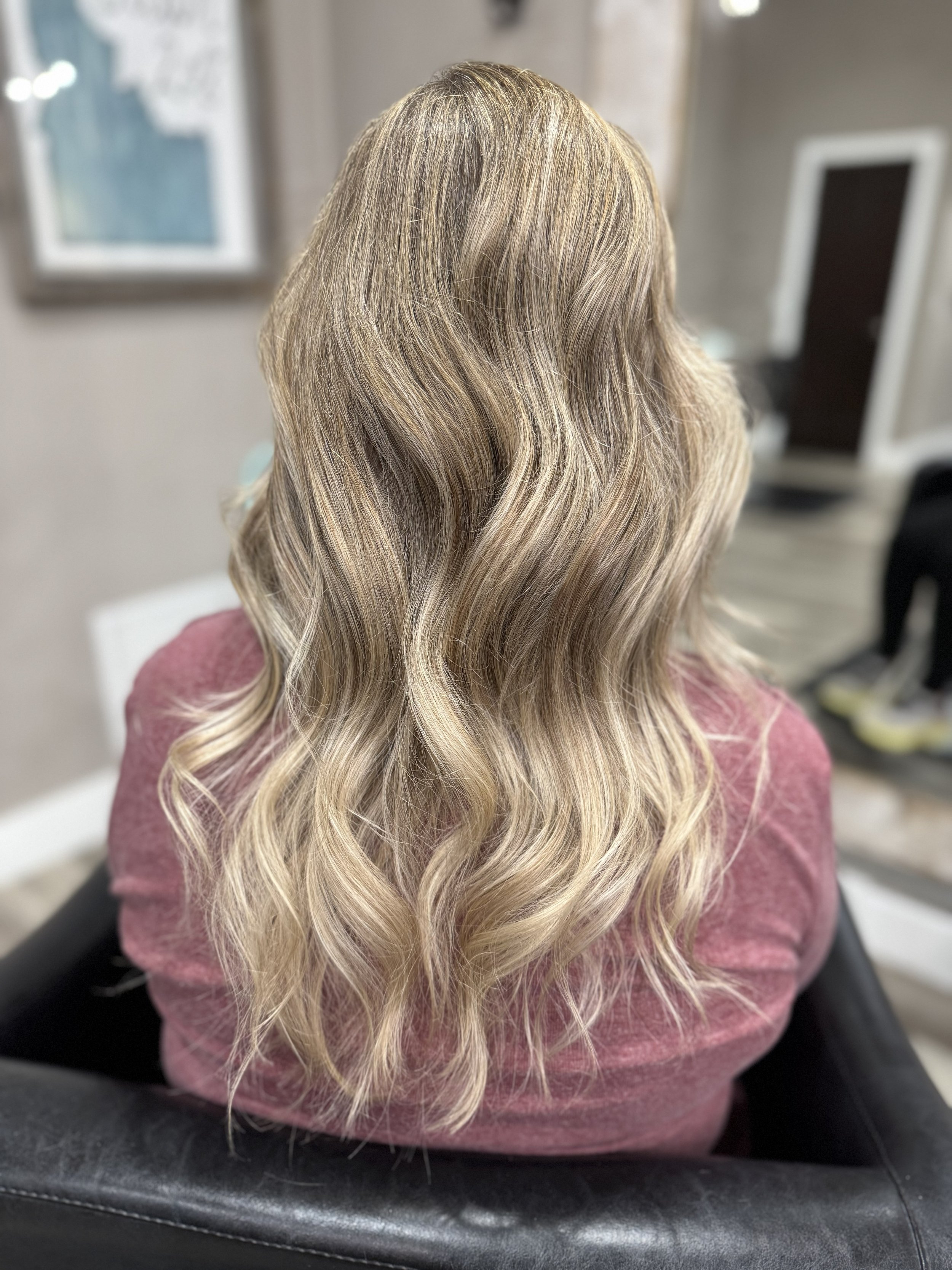 After the transformation, the back of a woman with long, wavy blonde hair wearing a pink top, sitting in a salon chair.