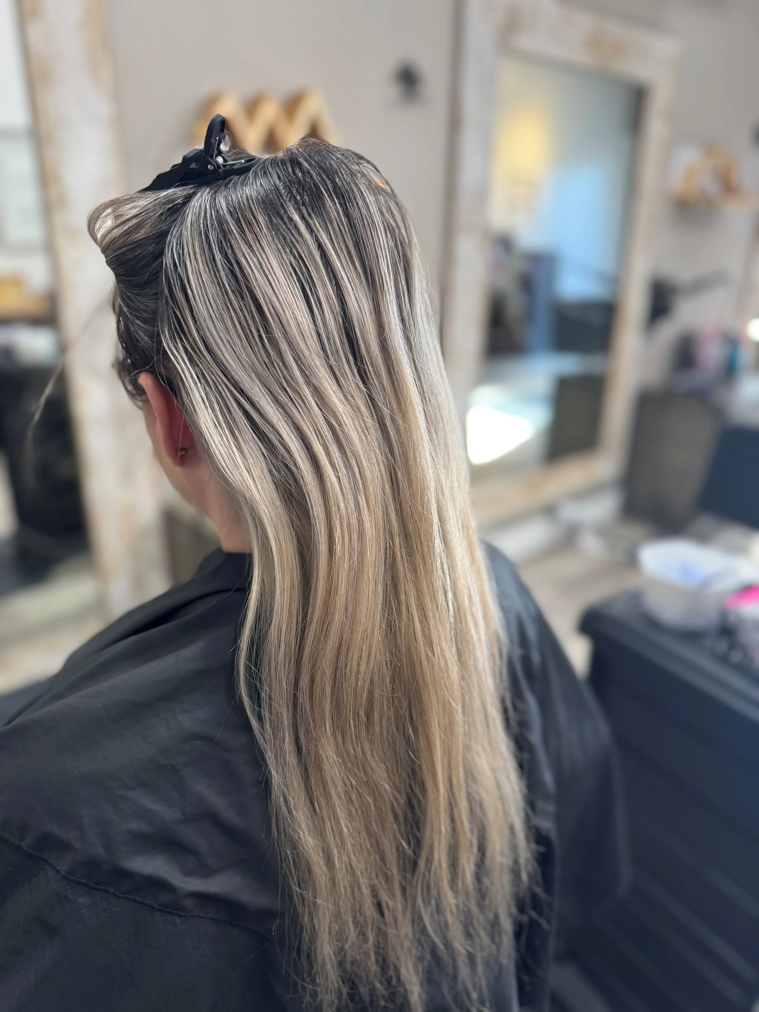 Back view of a woman with long, blonde hair getting her hair colored or styled at a salon.
