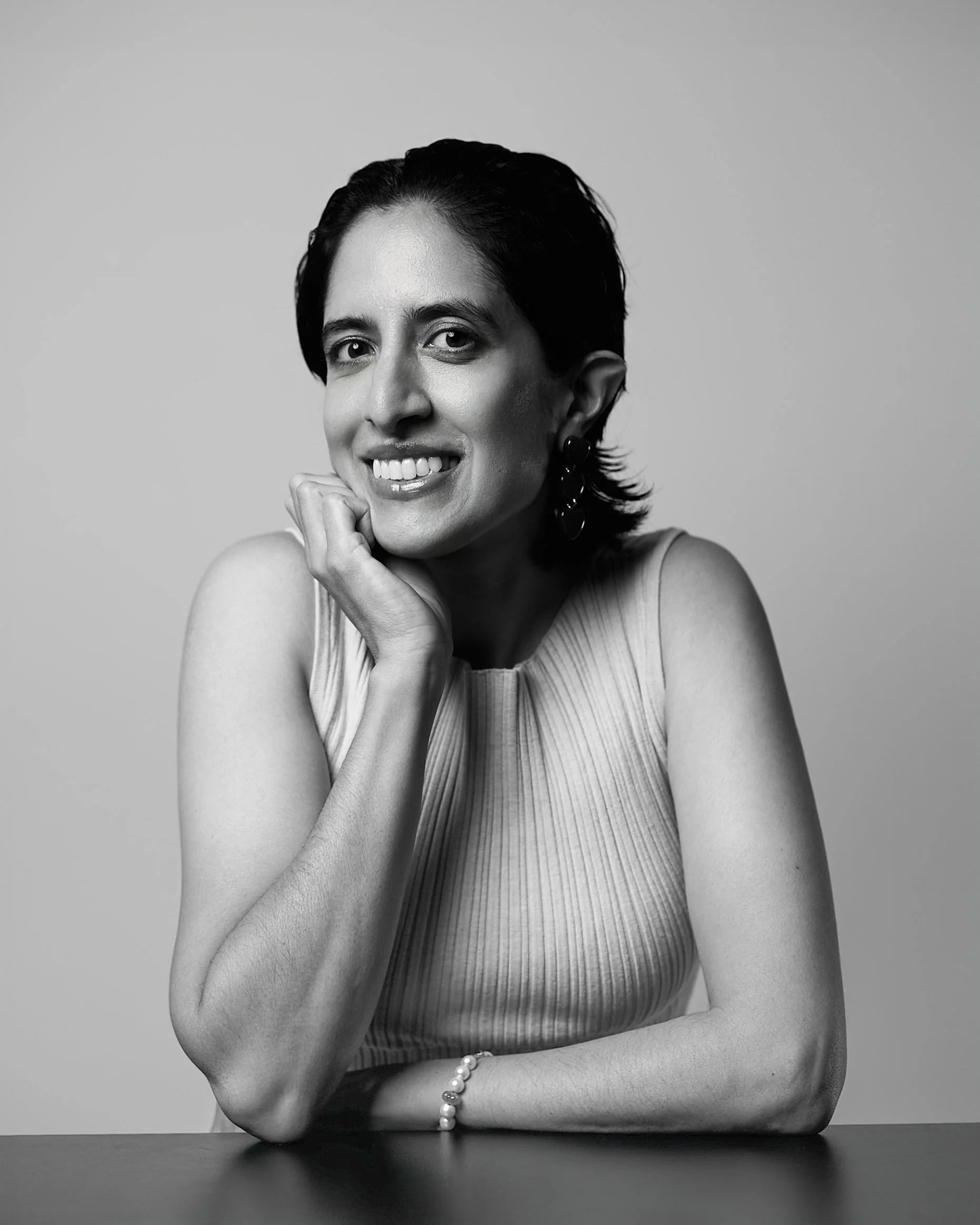 Black-and-white portrait of a woman with short dark hair, smiling and resting her chin on her hand, wearing a sleeveless striped top and a pearl bracelet.