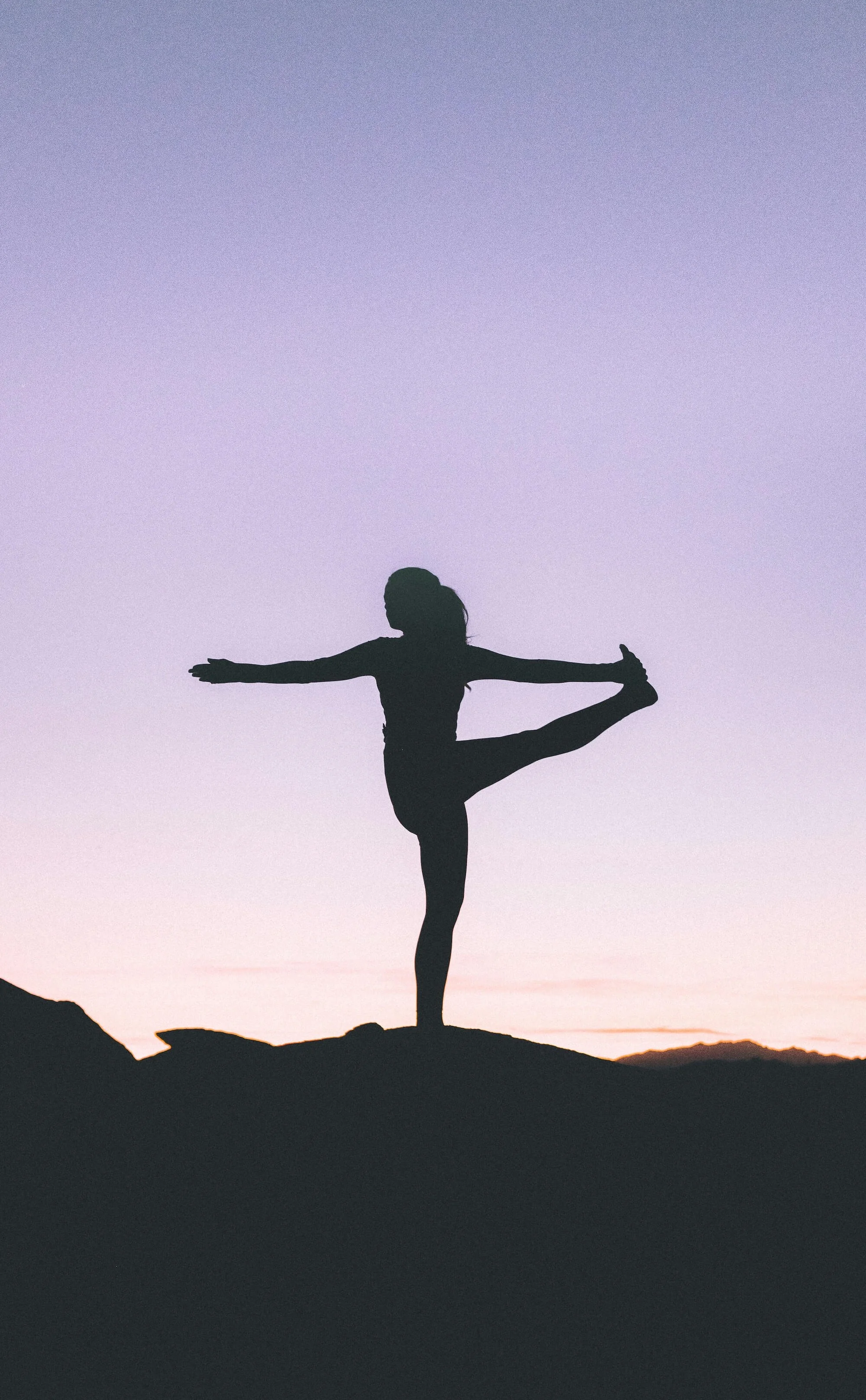 Silhouette of a person practicing yoga on a rocky hilltop during sunset, balancing on one leg with arms extended, against a colorful sky transitioning from purple to pink.