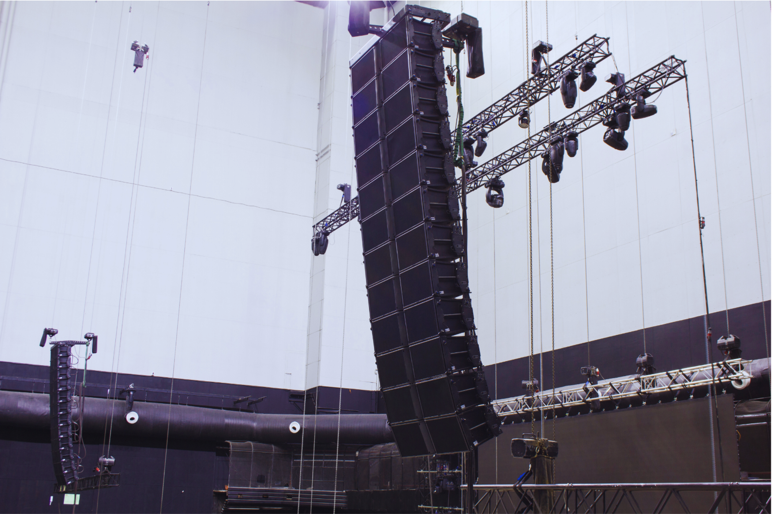 Large black speaker array suspended from metal trusses in an indoor venue, with additional stage lighting equipment.