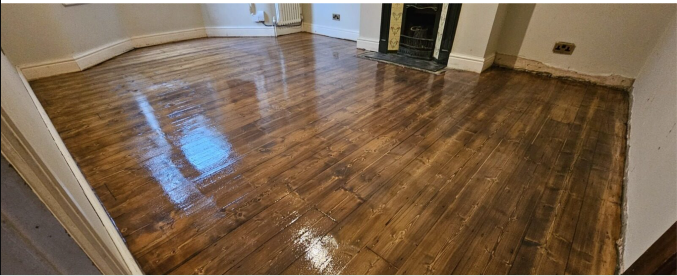 Photo of a freshly refinished hardwood floor in a living room with visible wood grain and a glossy finish, adjacent to a heater and an incomplete wall edge.