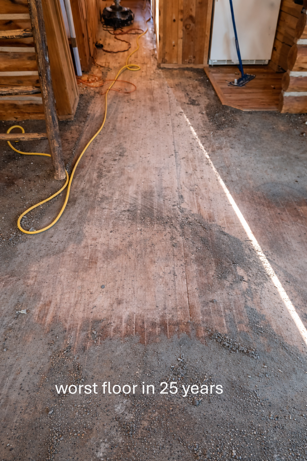 View of a room under renovation with a damaged, uneven wooden floor and visible dirt, debris, and dust. Construction tools and cords are present, indicating ongoing work.
