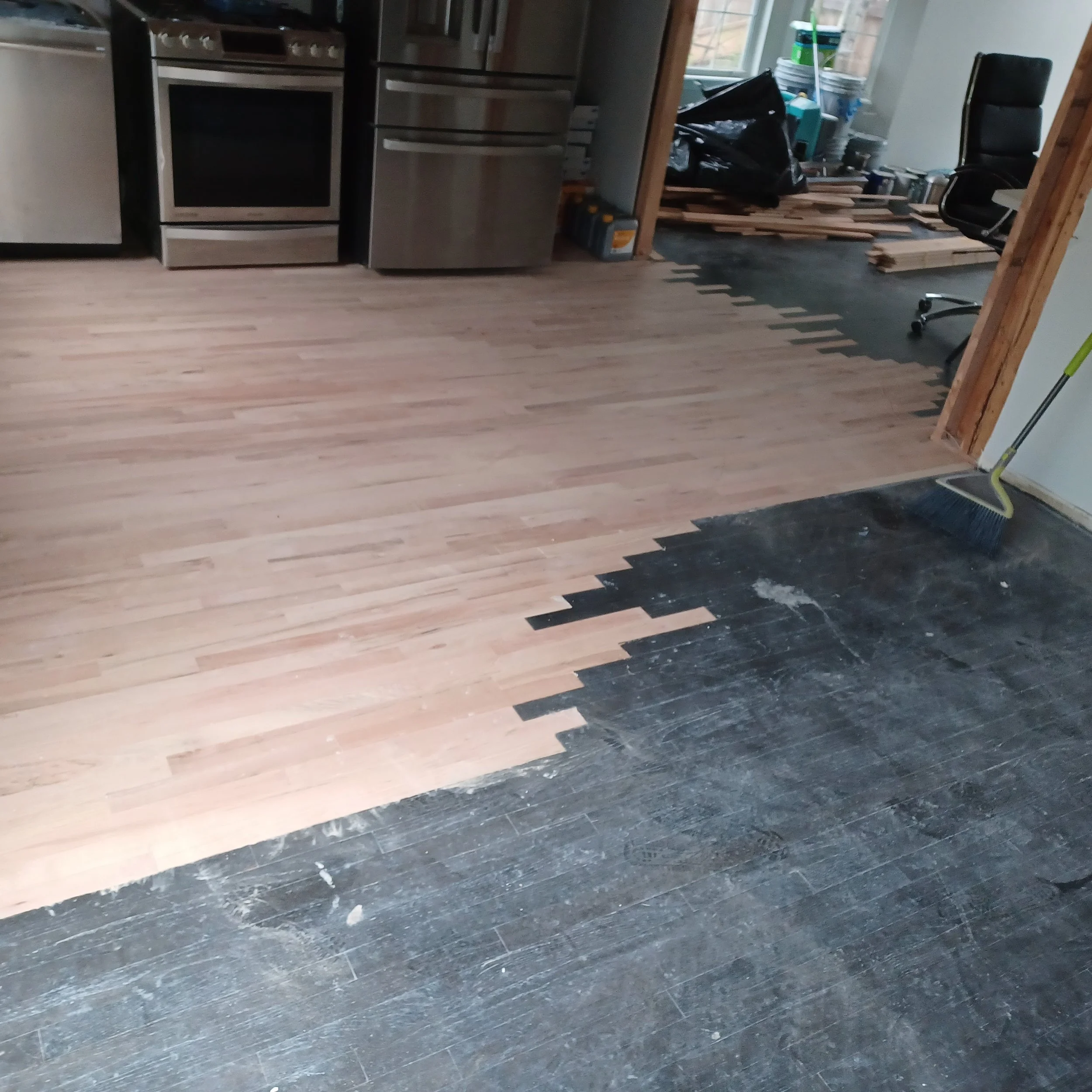 Partially installed hardwood flooring in a kitchen, with light-colored wood planks on one side and darker unfinished wood on the other, with construction tools and materials visible in the background.