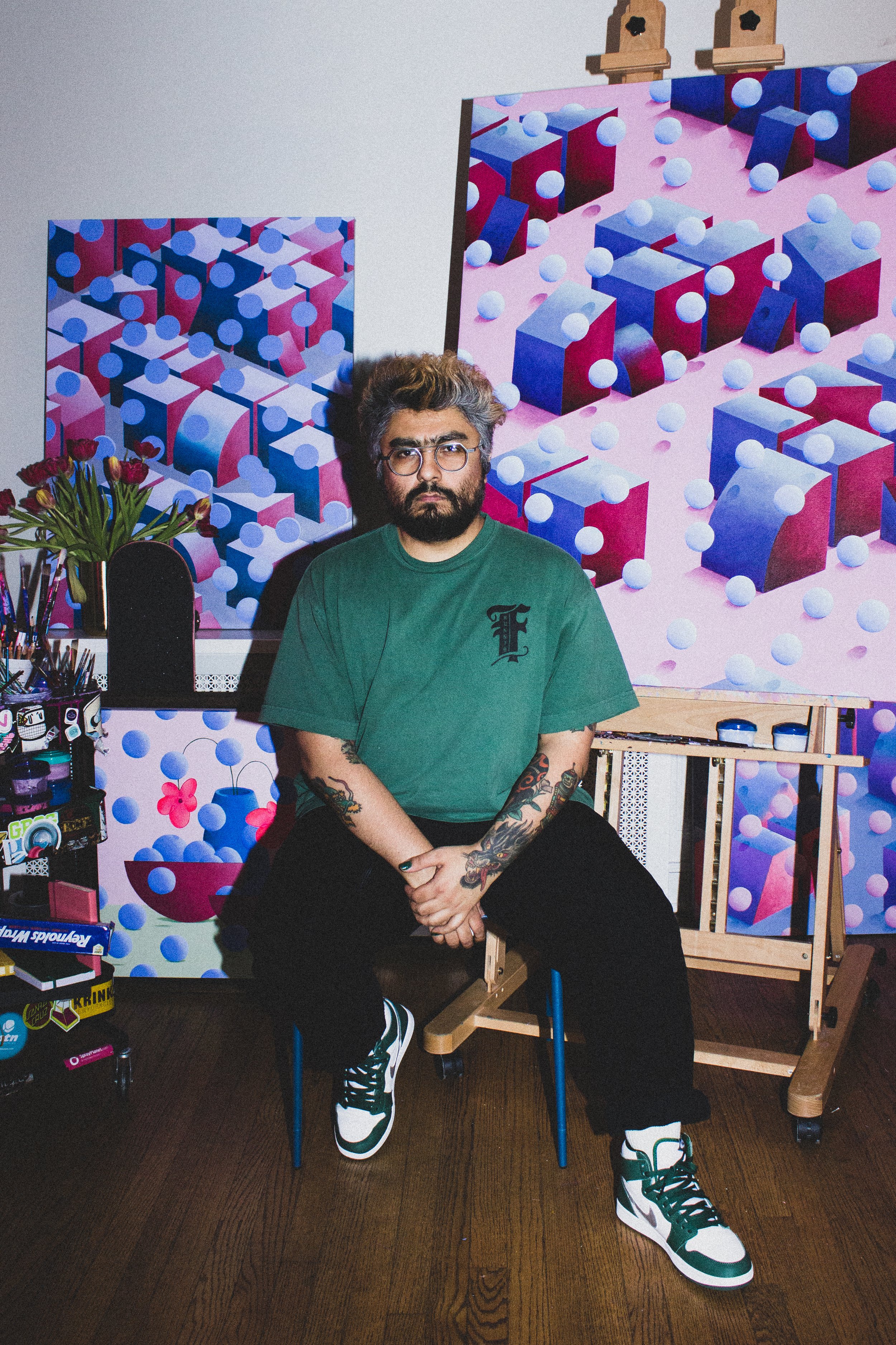 An artist with tattoos and glasses sitting in front of colorful abstract geometric paintings with pink, blue, and purple shapes and 3D spheres, in an art studio.