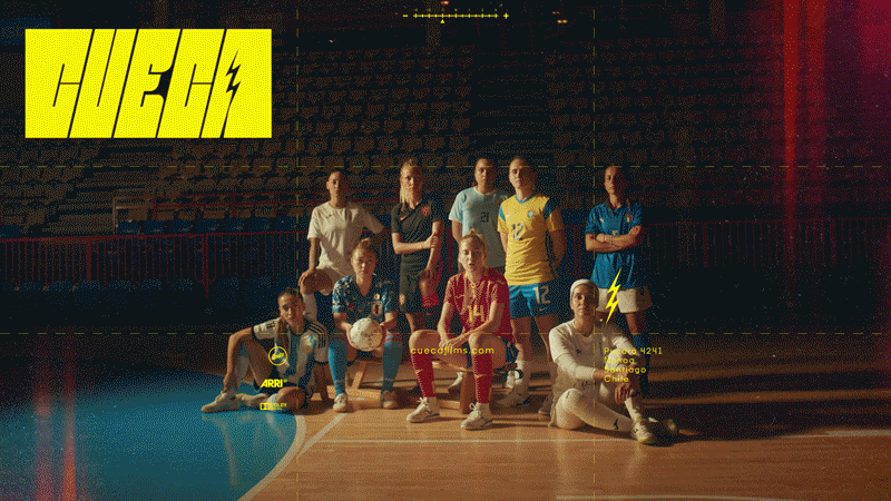 A group of female athletes in sports uniforms posed on an indoor basketball court, with chairs and empty seats in the background, and a yellow logo with a lightning bolt in the top left corner.