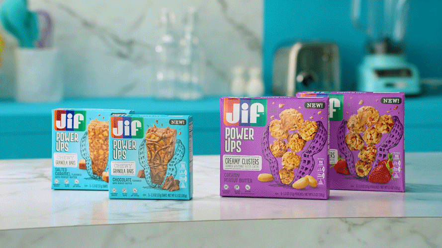 Four boxes of Jif Power Ups snack products on a white kitchen counter, with a light blue backsplash and kitchen appliances in the background. The boxes include Cherry Granola Bars, Chewy Salted Caramel Granola Bars, Creamy Clusters Peanut Butter, and Strawberry yogurt clusters.