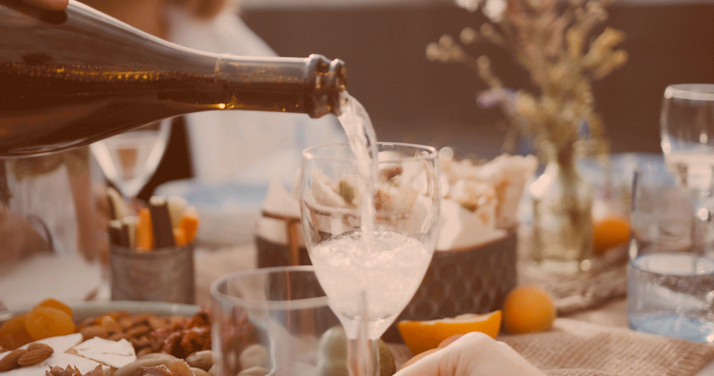 A person's hand is pouring water into a wine glass on a dinner table set with food and drinks.