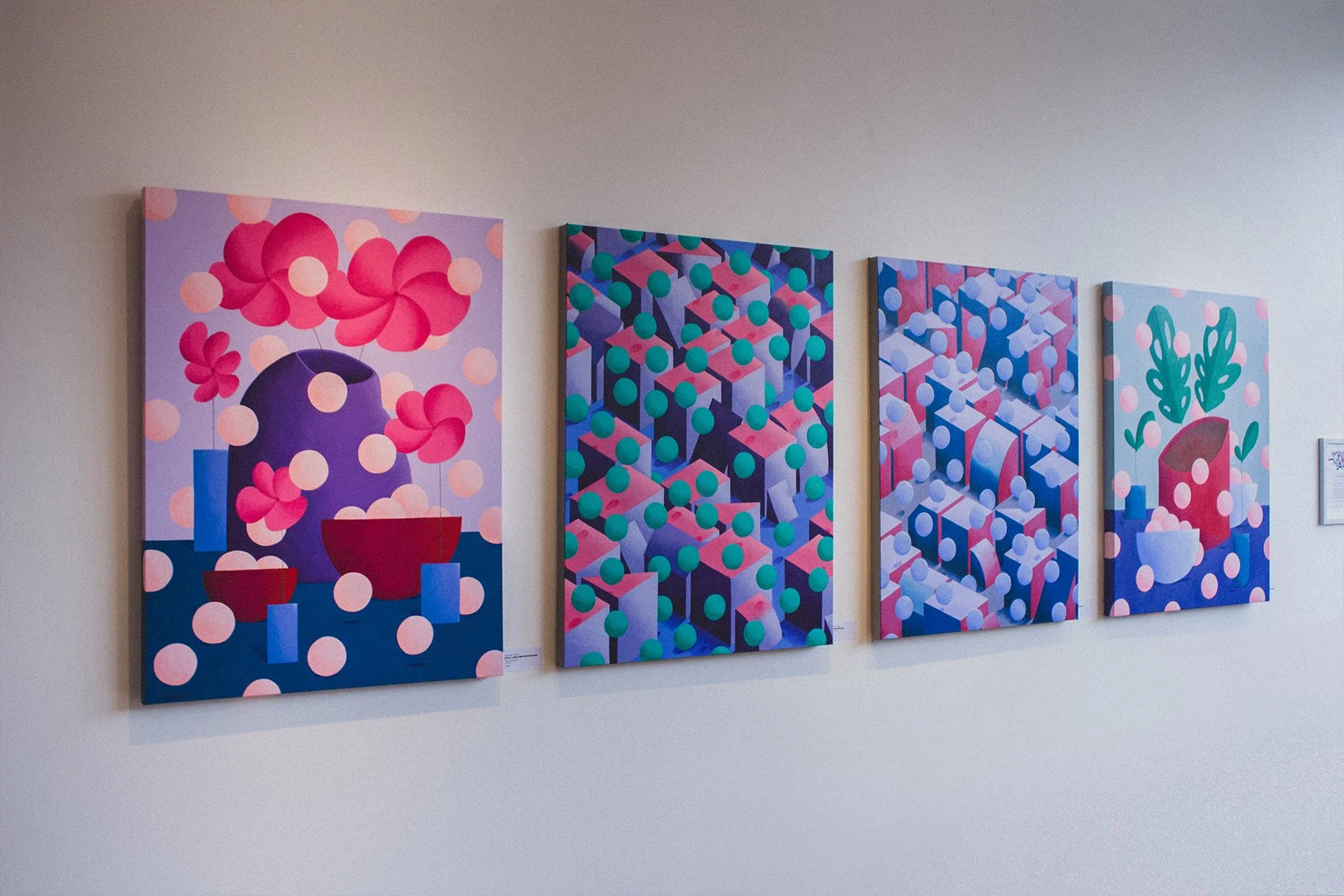 Four colorful, abstract paintings hanging on a white gallery wall, featuring geometric and floral patterns in shades of pink, purple, blue, and green.