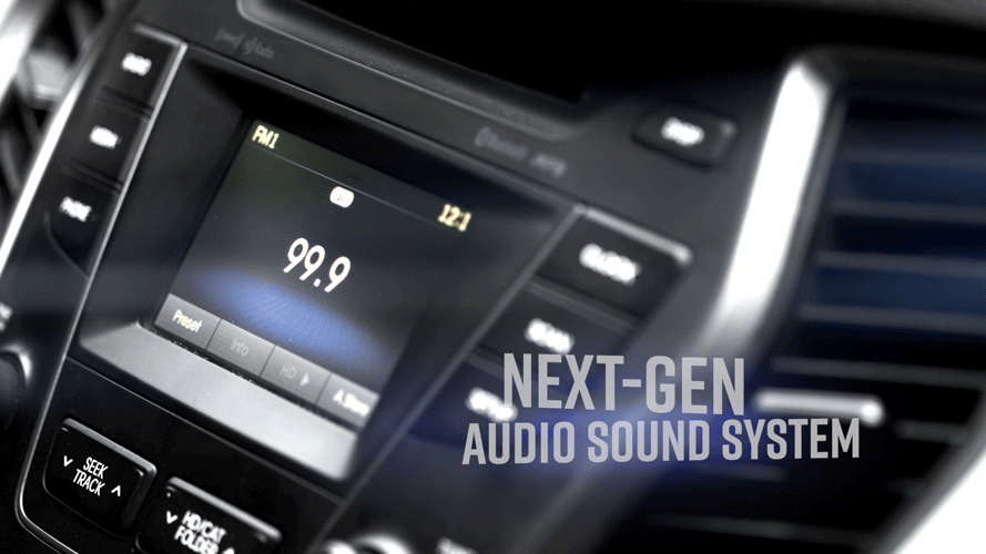 Close-up of a car stereo display showing 99.9 FM, with the text 'Next-Gen Audio Sound System' on the right side of the image.