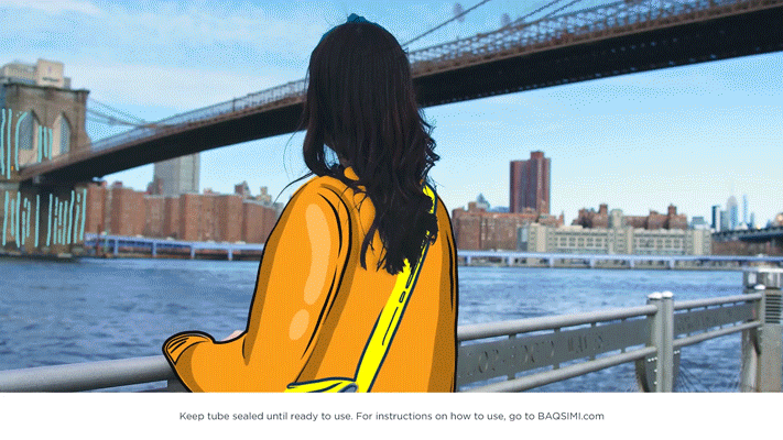 A woman with long dark hair wearing a yellow jacket and a bright yellow shoulder bag, standing by a waterfront with railing, looking towards a bridge over a river, with city buildings in the background.