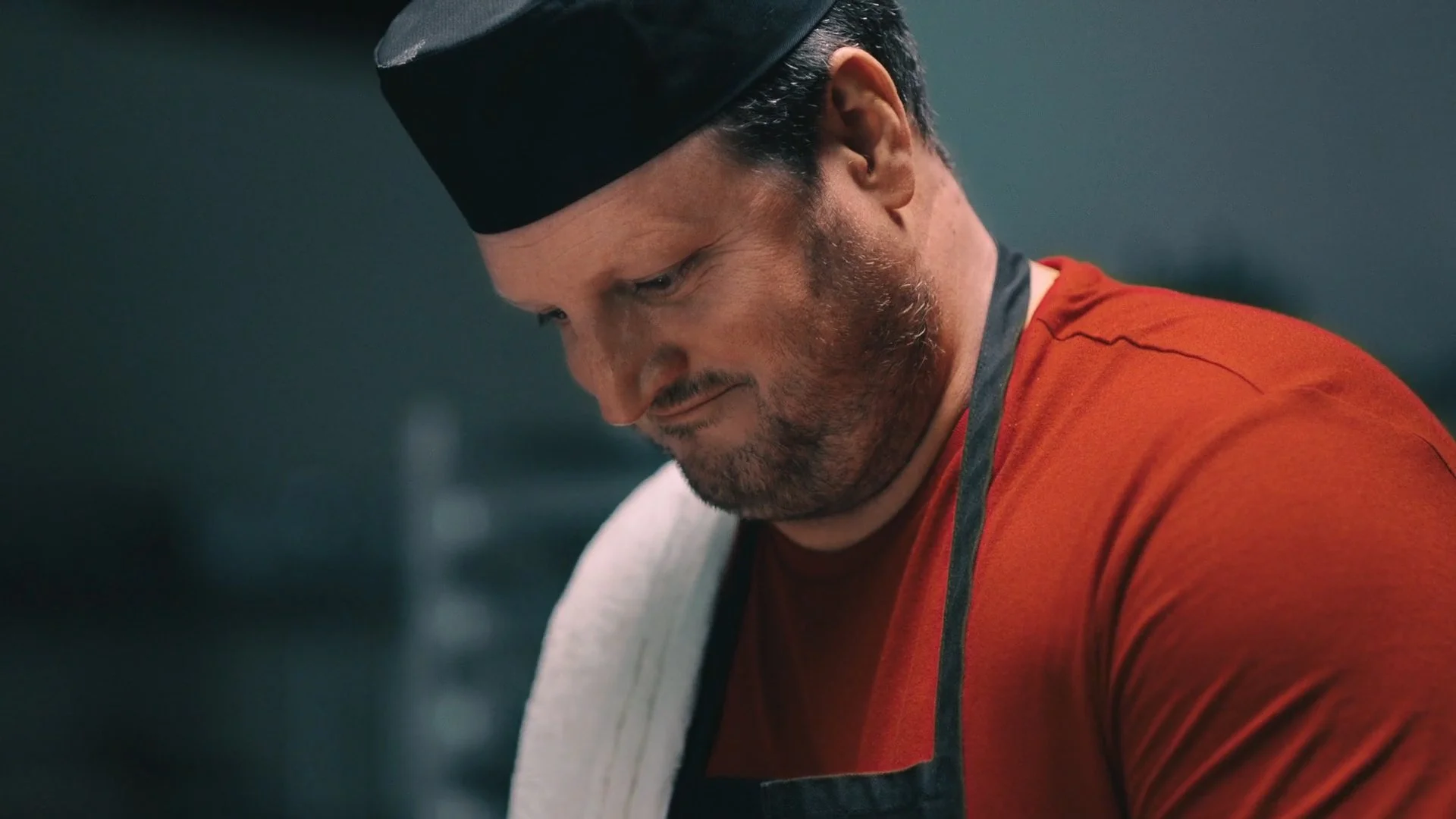 A man with a beard in a red T-shirt and black cap looks down thoughtfully with a white towel draped over his shoulder.